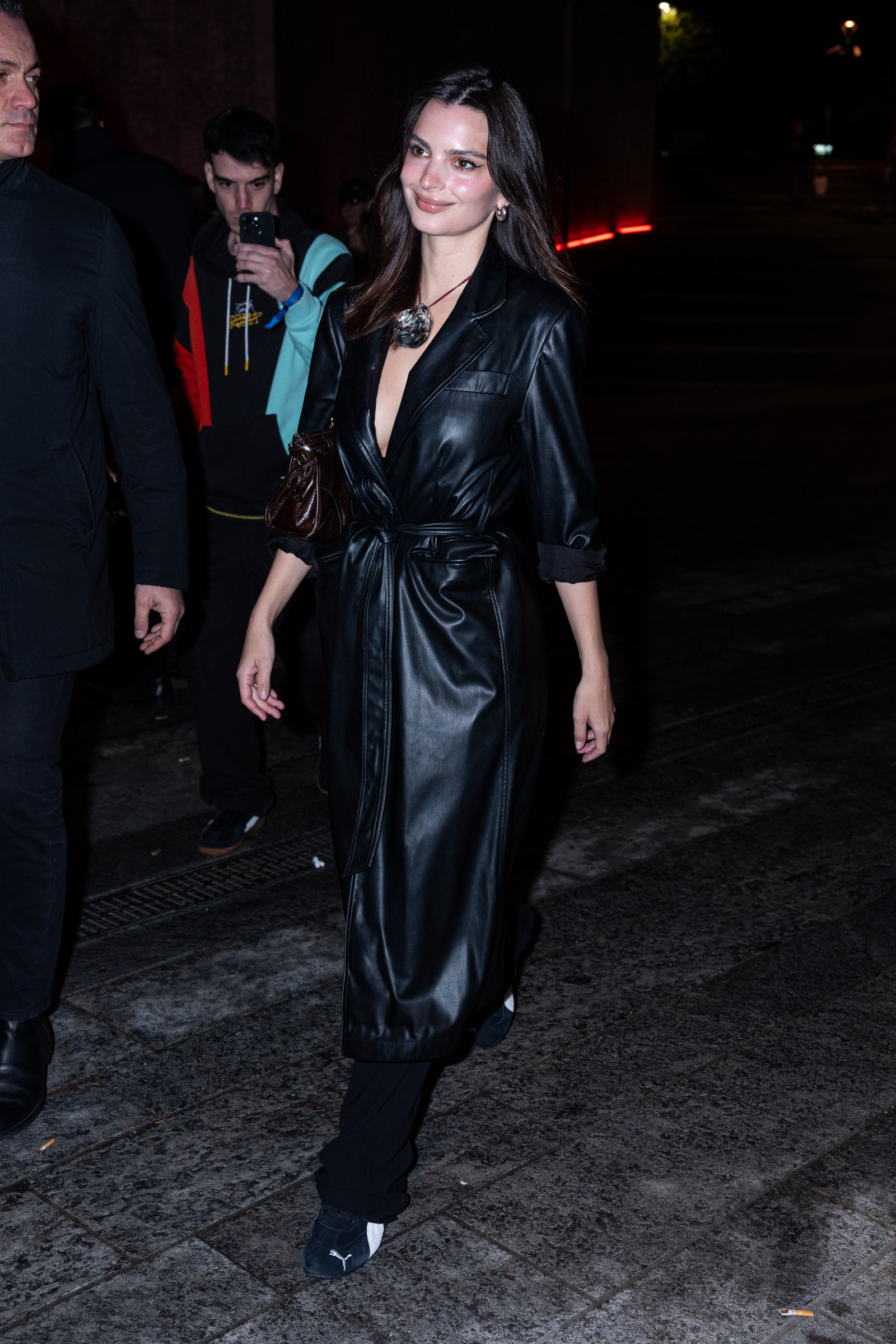 Emily Ratajkowski w czarnej skórzanej sukience i biało-czarnych sneakersach Puma Speedcat idzie w blasku ulicznych świateł.