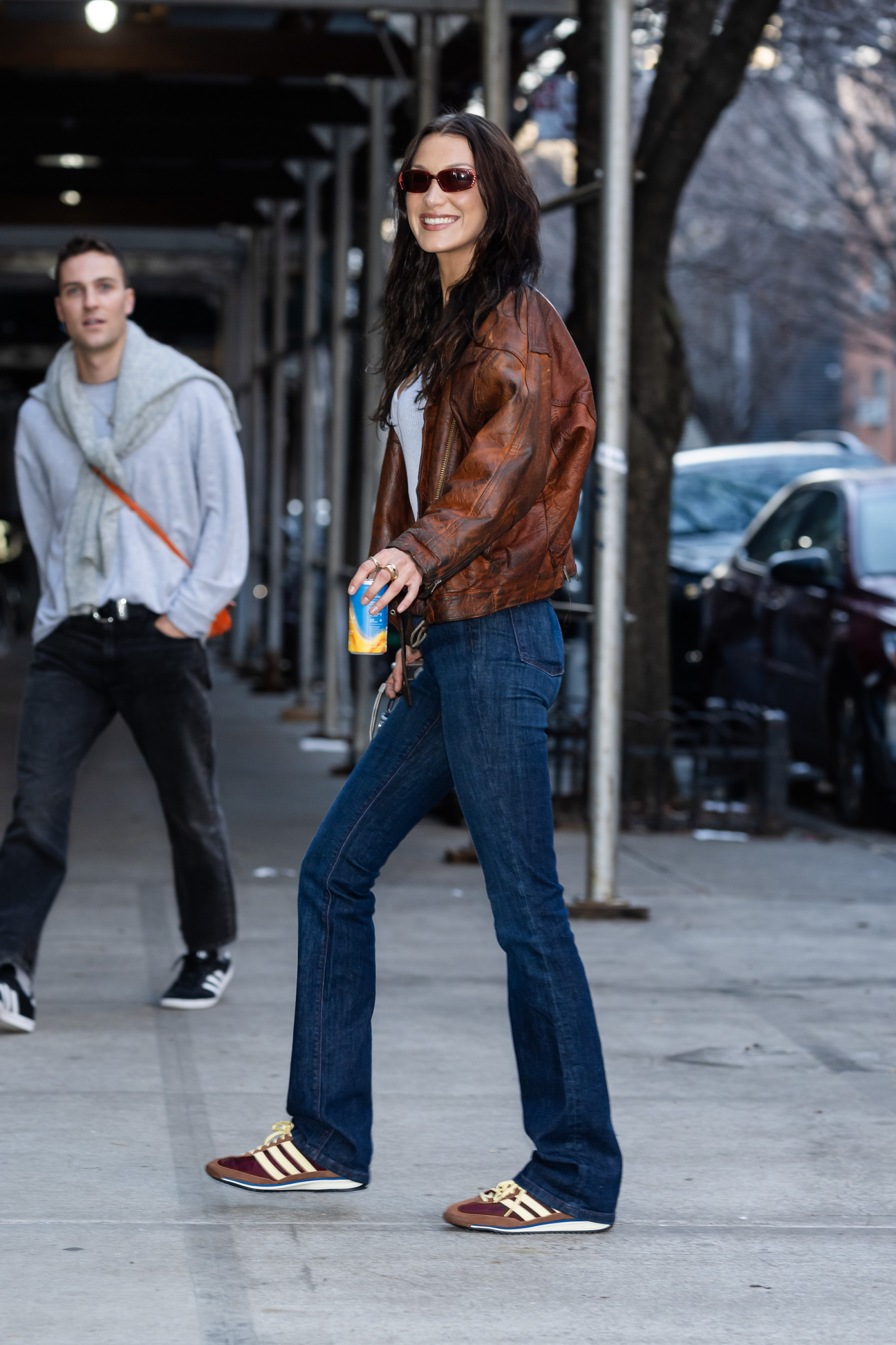 Bella Hadid w brązowych sneakersach adidas, w skórzanej kurtce, uśmiecha się na ulicy, trzymając napój.