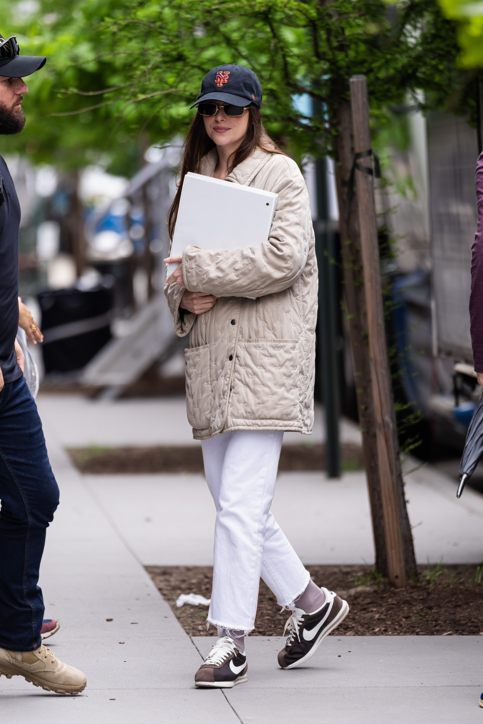 Dakota Johnson w brązowych sneakersach Nike Cortez, niesie laptopa i nosi jasną kurtkę w miejskim otoczeniu.