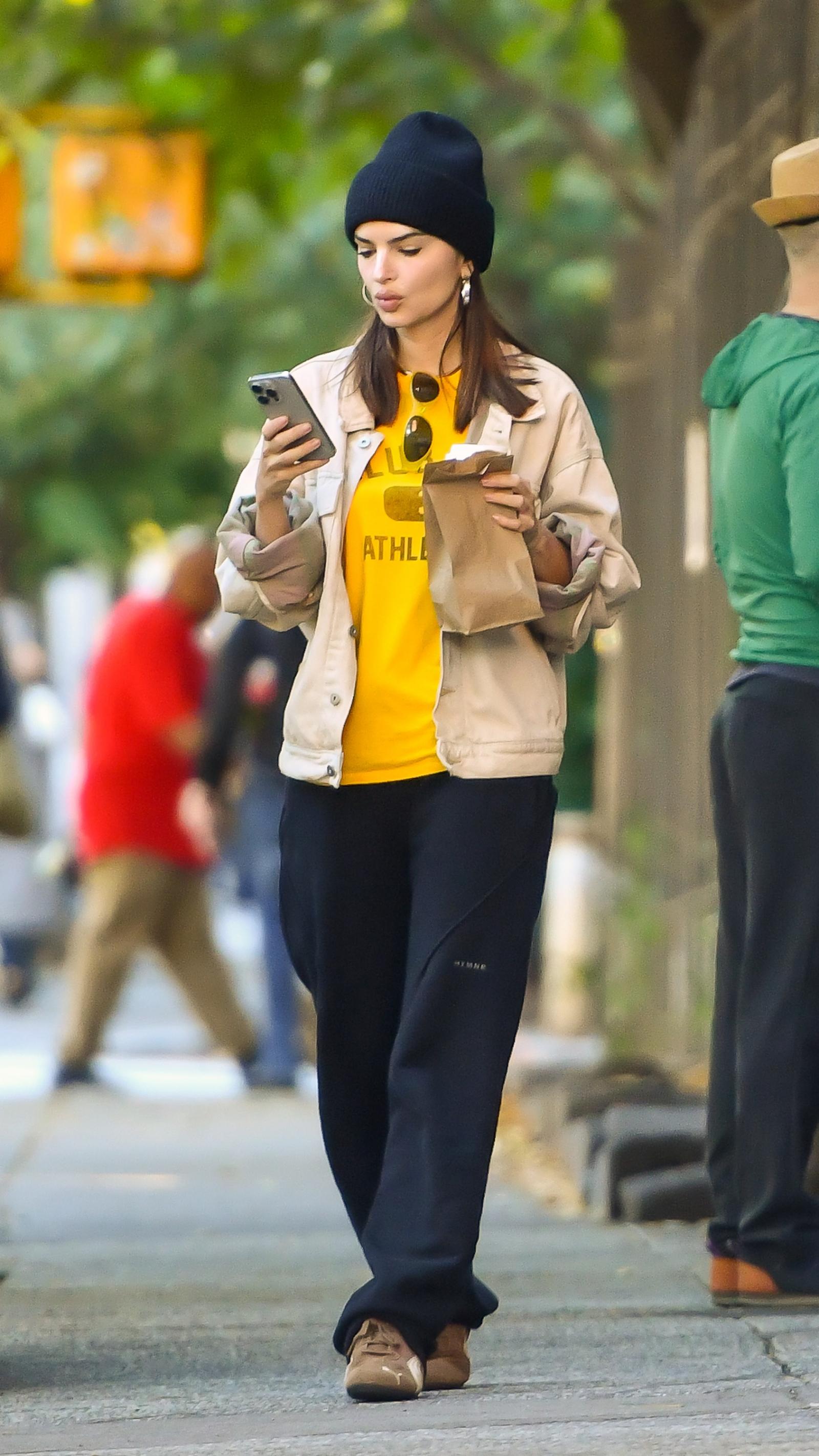 Emily Ratajkowski w brązowych sneakersach Puma, w jasnej kurtce i żółtej koszulce, sprawdza telefon na ulicy.