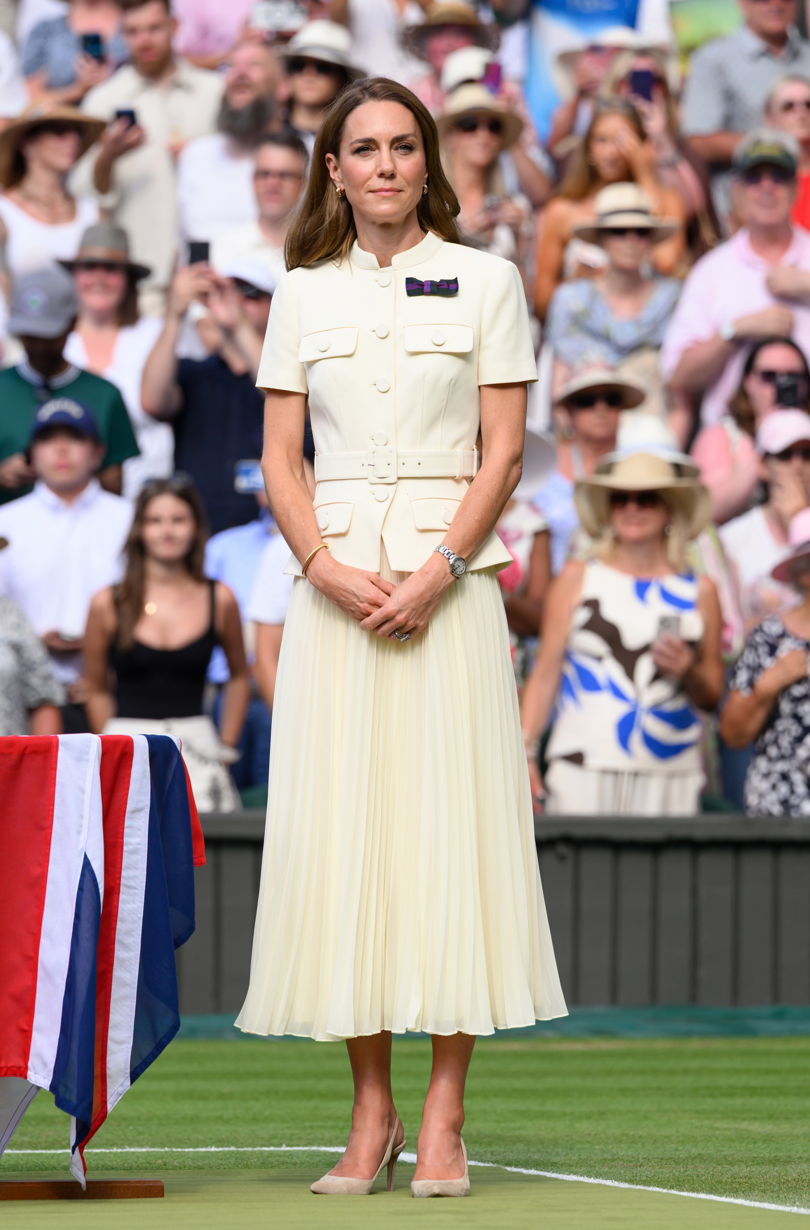 Kate Middleton na Wimbledonie, ubrana w długą maślaną spódnicę plisowaną, stoi wśród tłumu widzów.