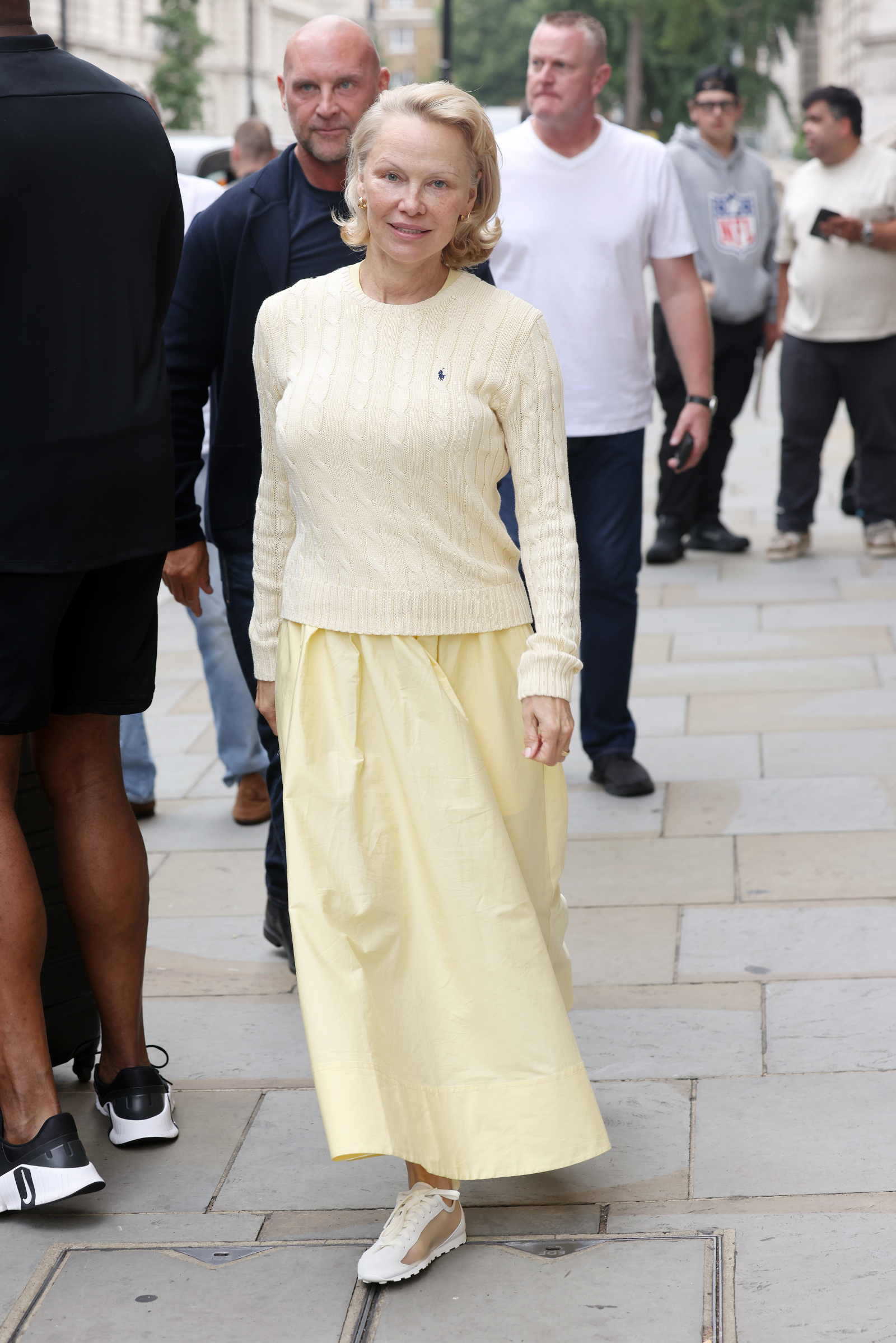 Pamela Anderson w spódnicy maxi w kolorze yellow butter i swetrze, spacerująca w miejskim otoczeniu.