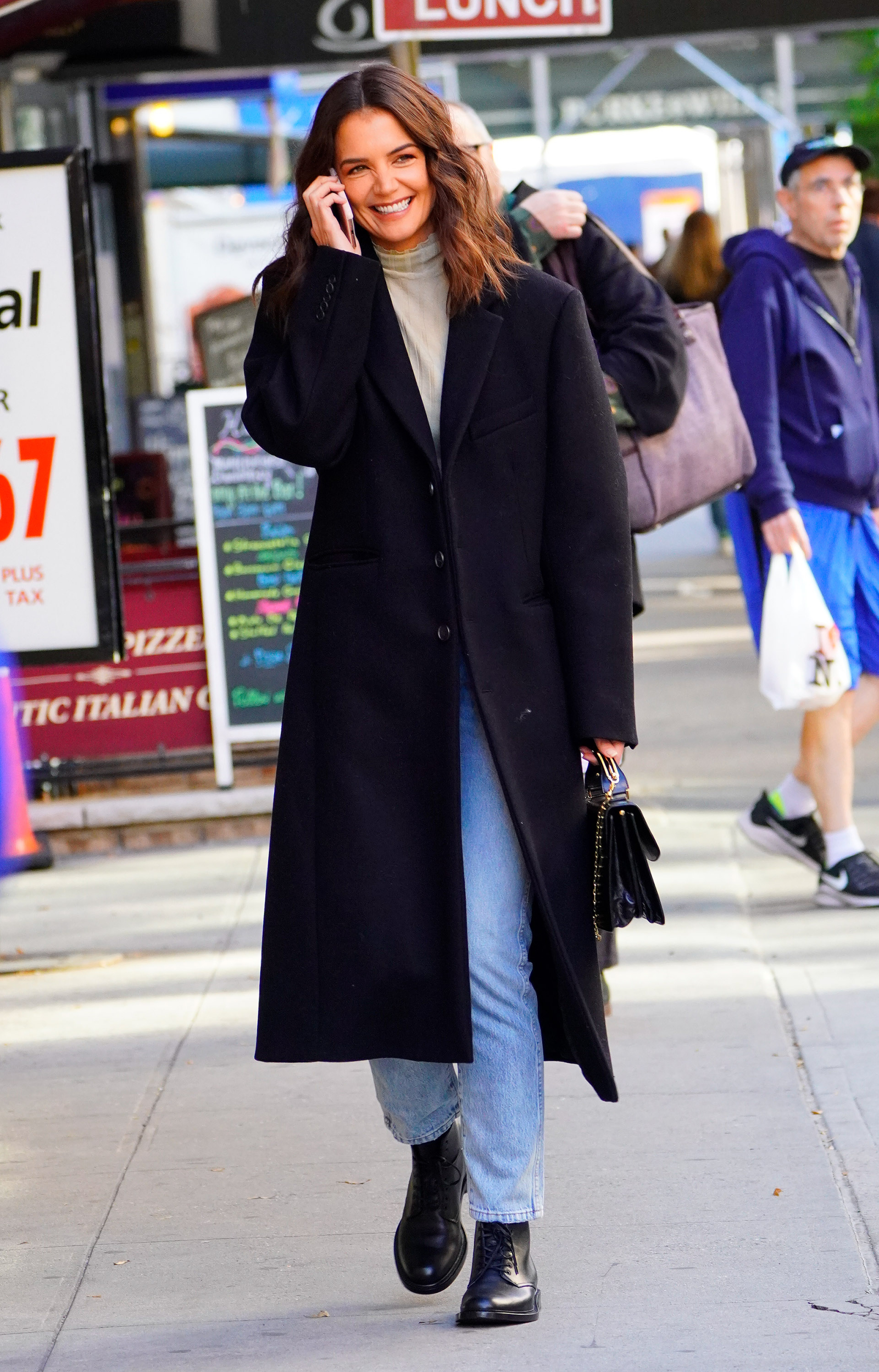 Katie Holmes w prostych jeansach i dopasowanym golfie, uśmiechając się, rozmawia przez telefon na ulicy.