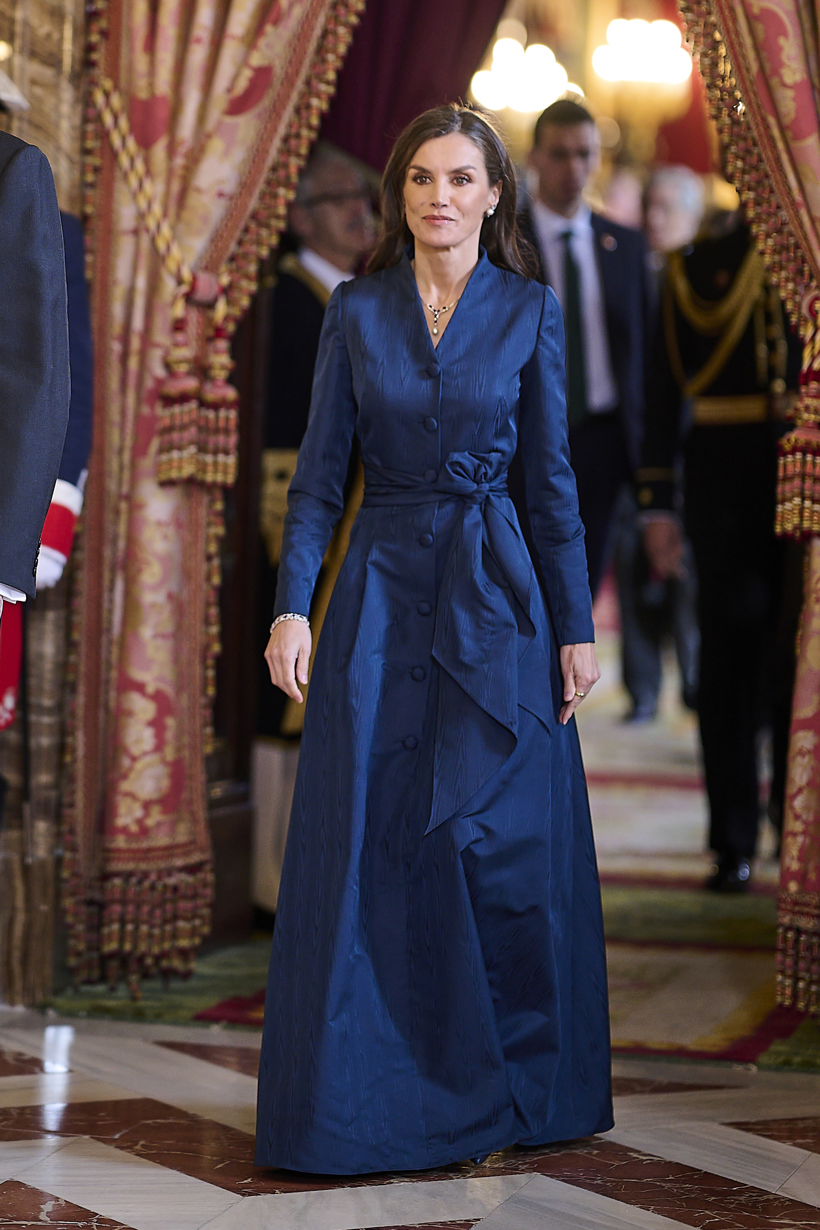 Królowa Letycja w granatowej sukni maxi, elegancko spacerująca po reprezentacyjnym pomieszczeniu.