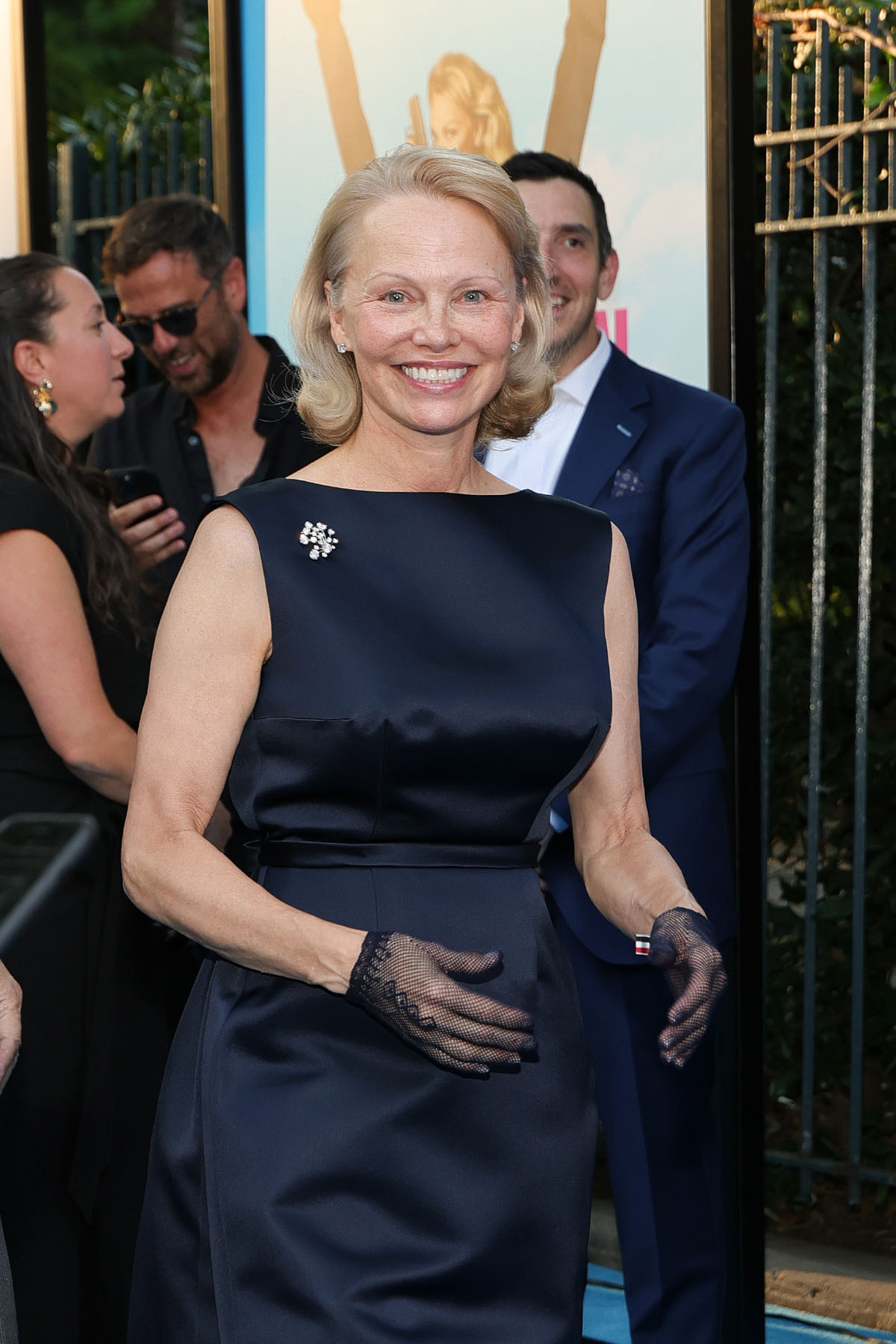 Pamela Anderson w granatowej sukni maxi z rękawiczkami, uśmiechnięta na tle imprezy.