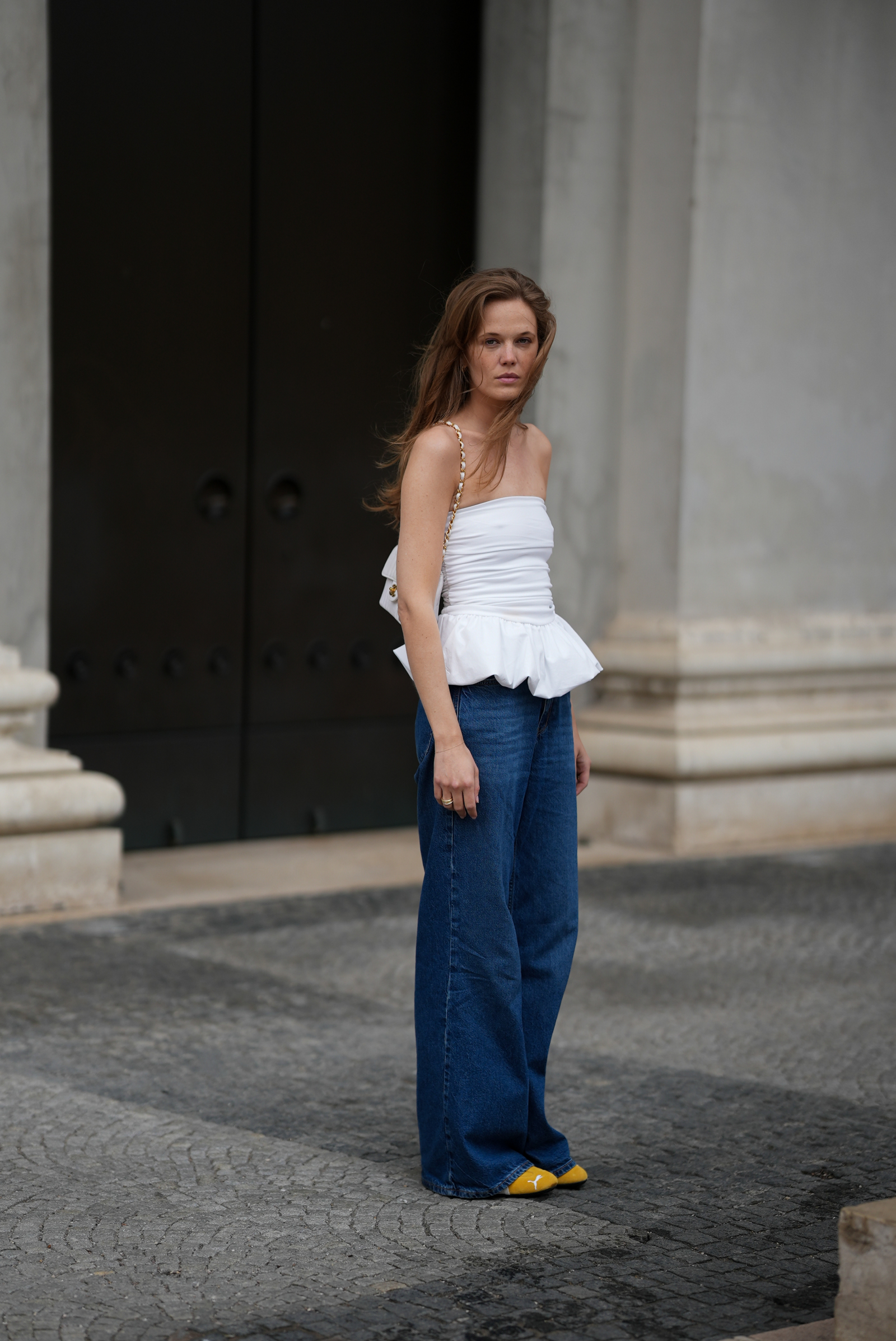 Kobieta w topie z baskinką i baggy jeans, stoi na kamiennej nawierzchni przed dużymi drzwiami.