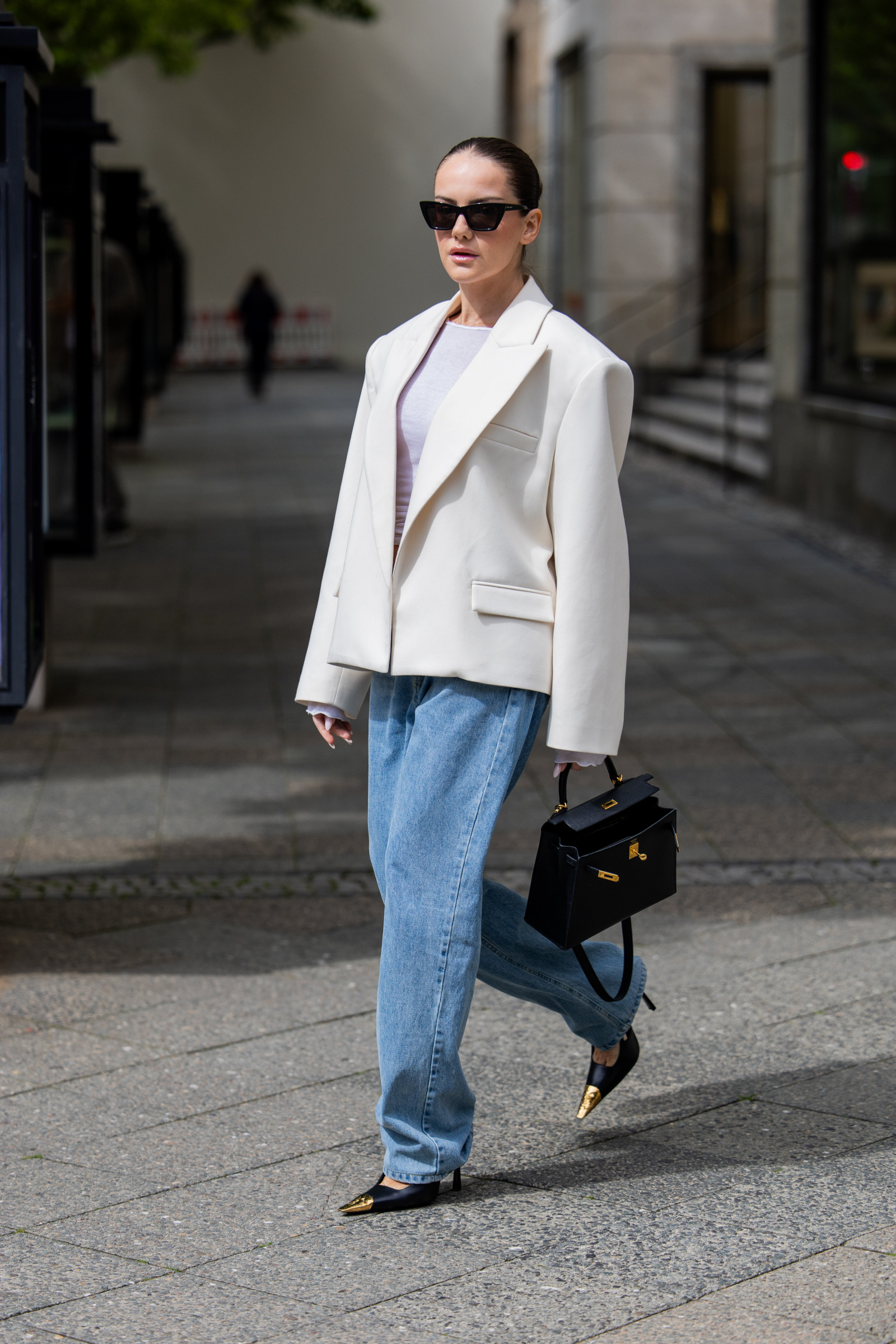 Kobieta w białej marynarce, baggy jeans i szpilkach z ostrym noskiem spaceruje po malowniczej ulicy.