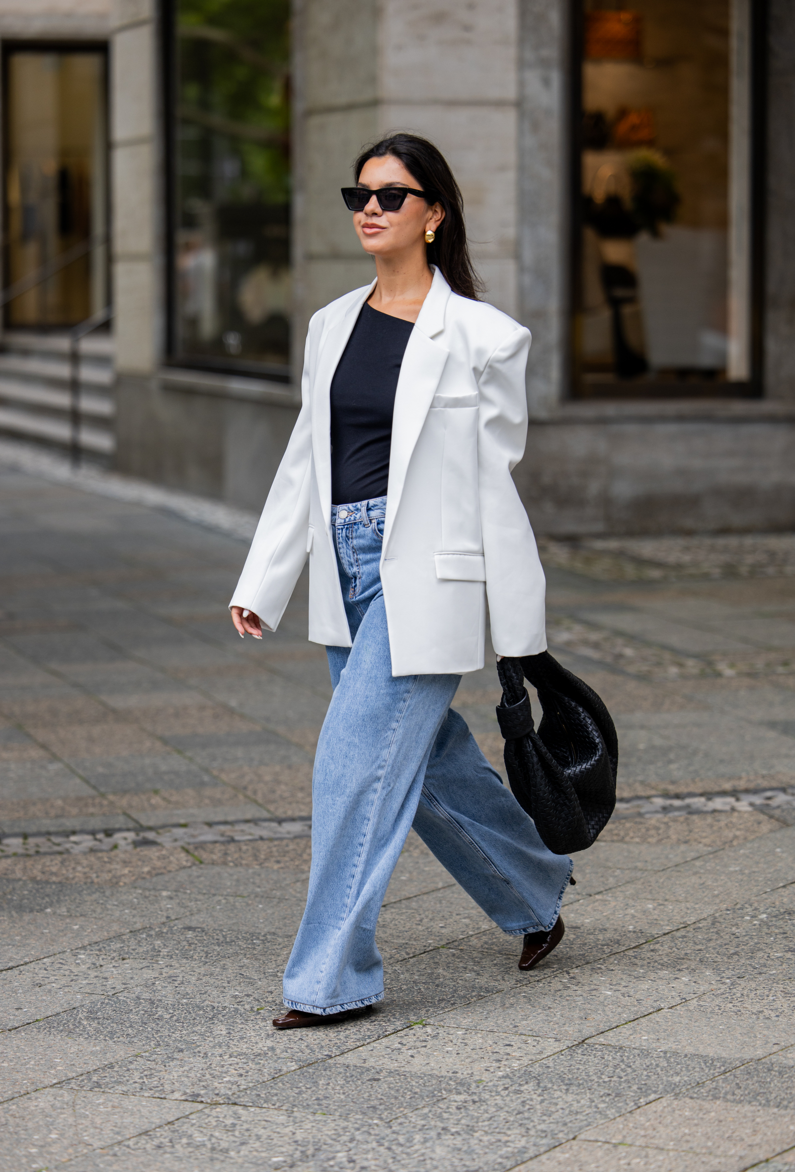 Kobieta w białej marynarce, baggy jeans oraz szpilkach z ostrym noskiem idzie po ulicy.