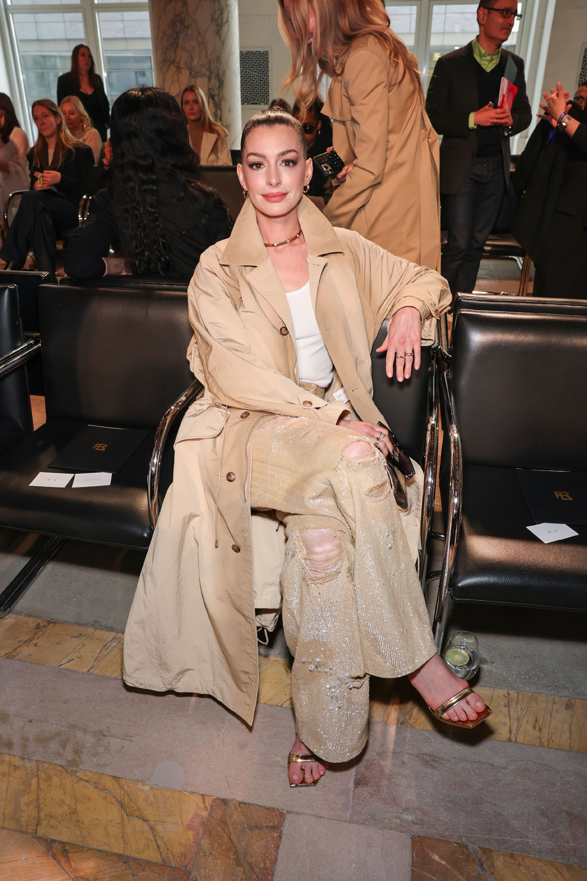Anne Hathaway w beżowym płaszczu, w sandałkach, bez pedicure, w bluzie Ralph Lauren, siedzi na pokazie.