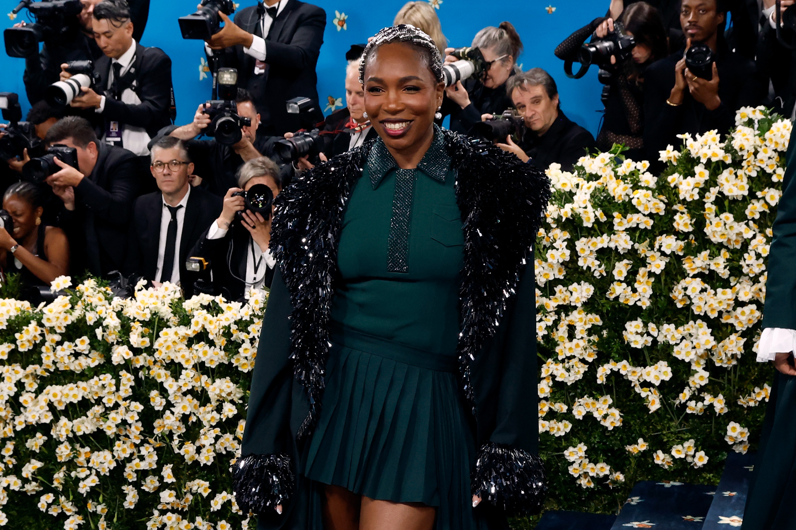 Venus Williams na Met Gala 2025, ubrana w elegancką sukienkę z ozdobnym płaszczem, w otoczeniu fotograf&oacute;w i kwiat&oacute;w.