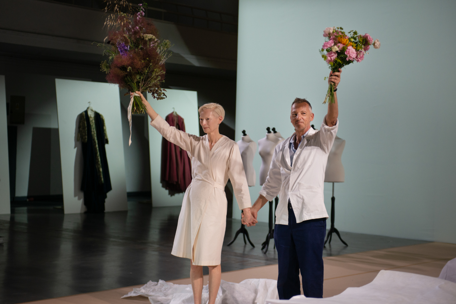 Tilda Swinton na Malta Festival 2025 prezentuje spektakl "Embodying Passolini" trzymając bukiety kwiatów.