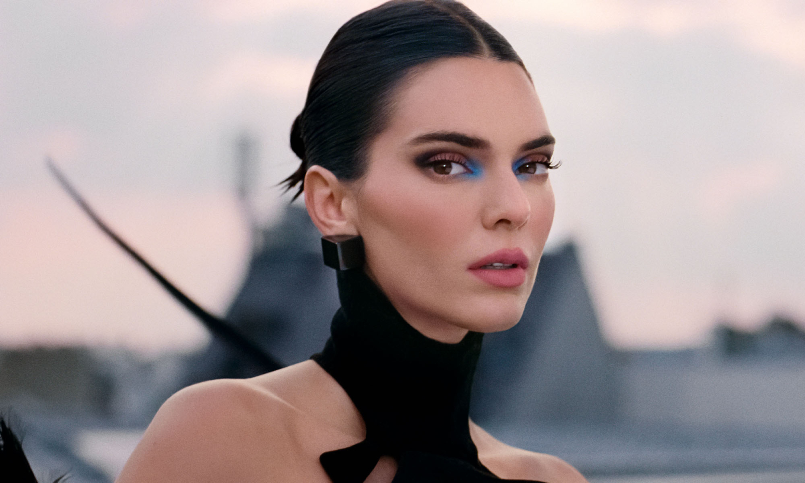 Kendall Jenner w eleganckiej kreacji z widocznym makijażem i ciekawą fryzurą, pozująca na tle miejskiego krajobrazu.
