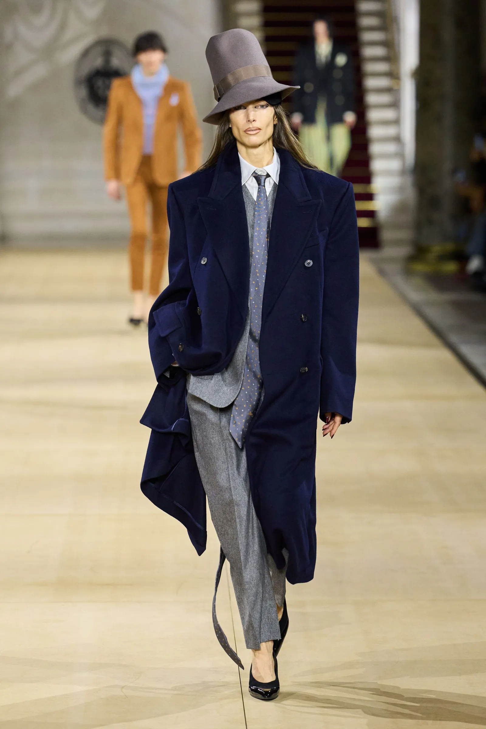 Modelka w granatowym płaszczu na zimę 2025 z szerokim kapeluszem i eleganckim zestawem harmonijnie przechodzi po wybiegu.