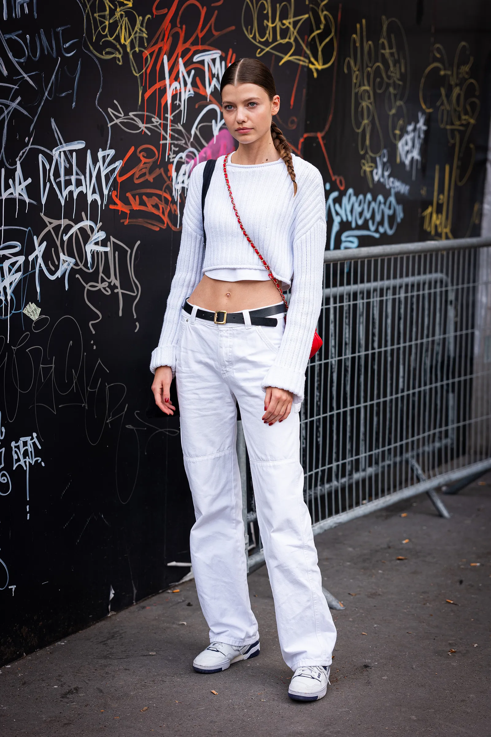 Modelka w białych jeansach i sneakersach, w białym swetrze, stoi przed graffiti na czarnym tle.