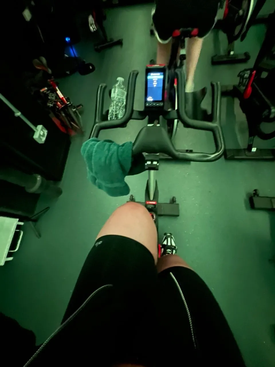 Widok z perspektywy osoby na rowerze stacjonarnym w fitness klubie. Widać zegar treningowy, wodę i ręcznik.
