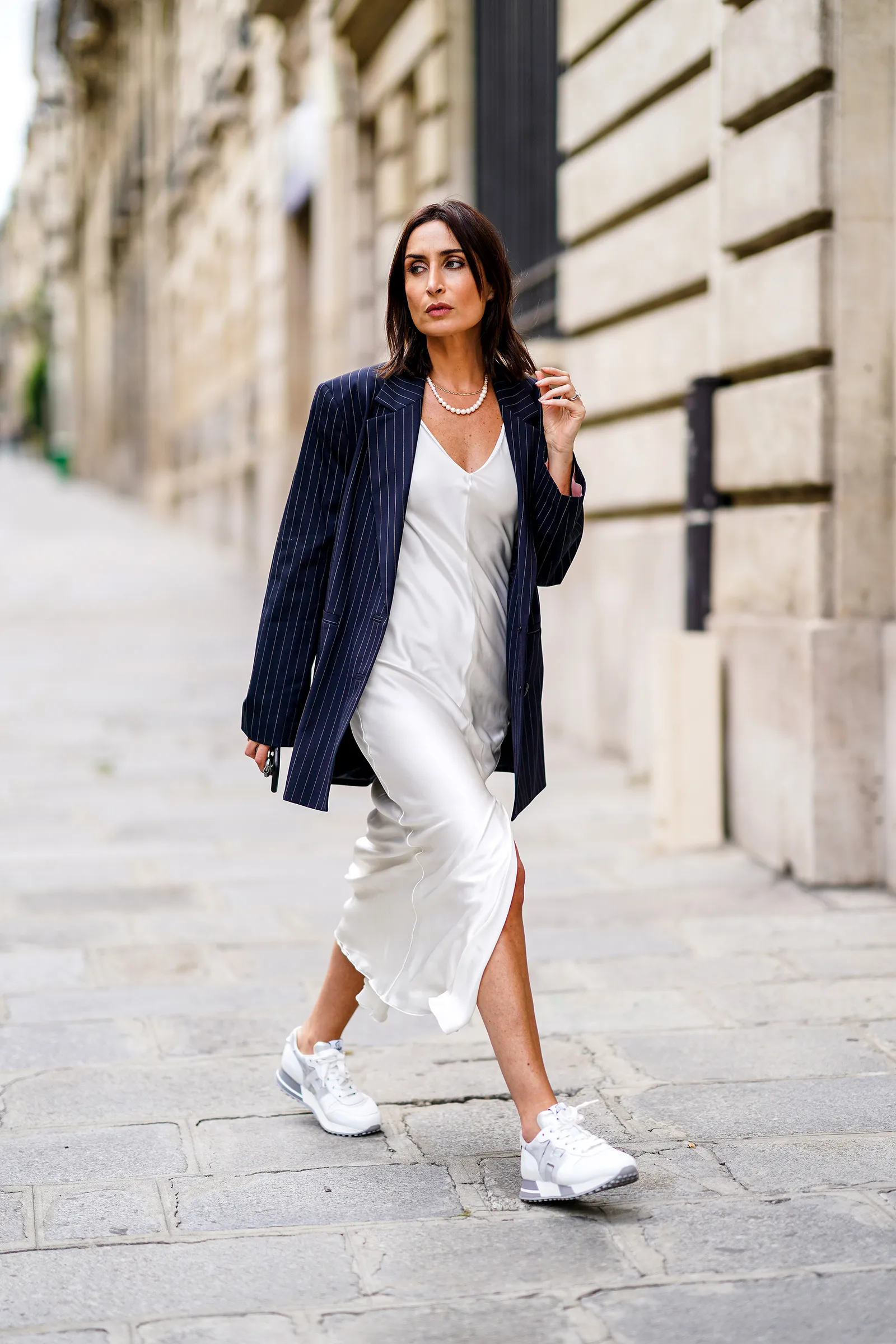 Kobieta w białej slip dress i stylowej marynarce spaceruje w sneakersach po ulicy.