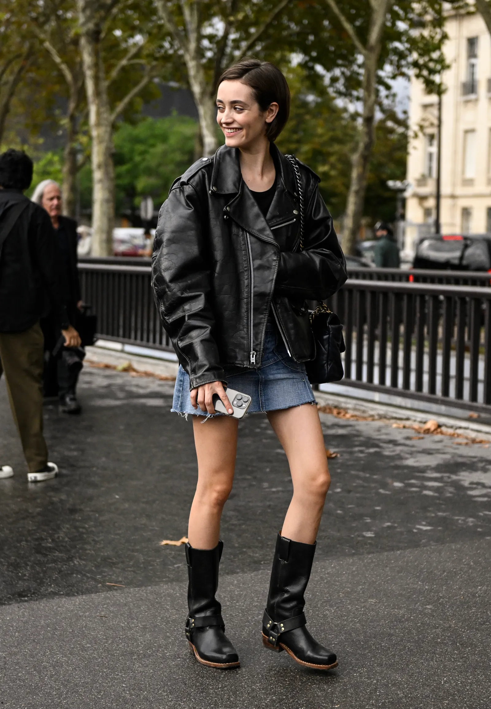 Młoda kobieta w skórzanej kurtce i dżinsowej spódnicy, nosi biker boots i uśmiecha się na ulicy.