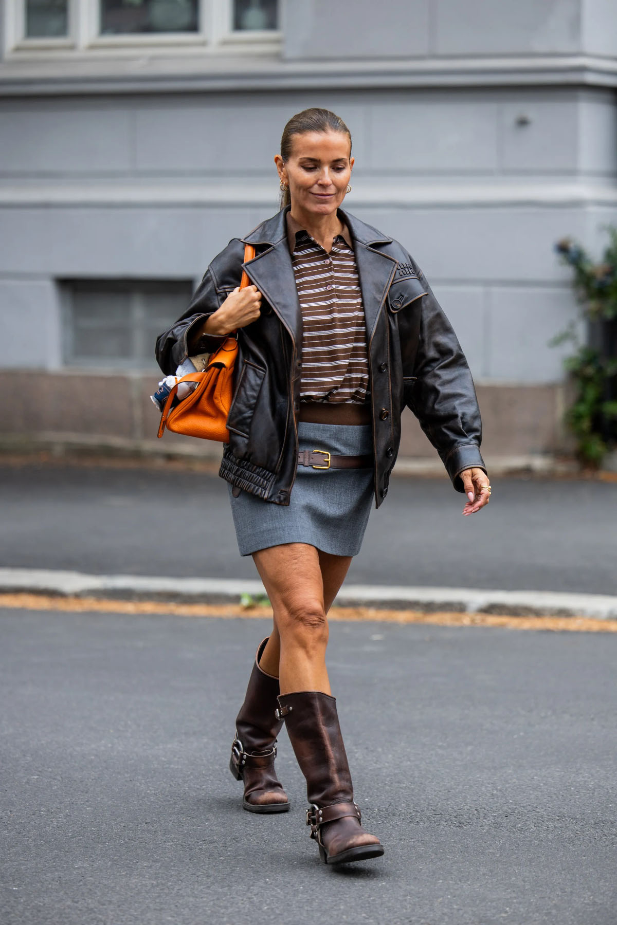 Kobieta w skórzanej kurtce i spódnicy, w biker boots, spaceruje ulicą z torbą w dłoni.