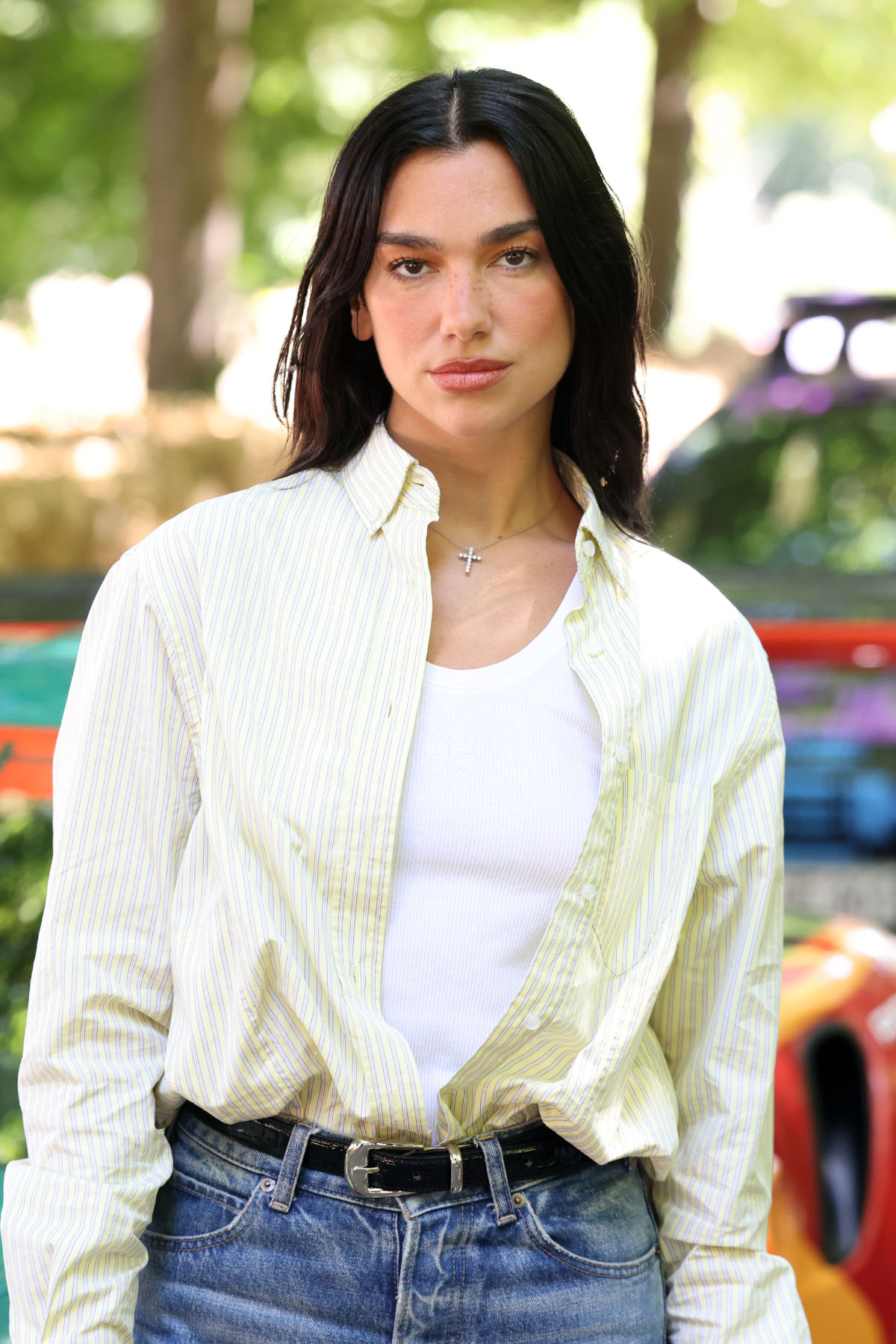 Dua Lipa w jasnej koszuli z naszyjnikiem z krzyżem, pozująca na tle kolorowych pojazdów.