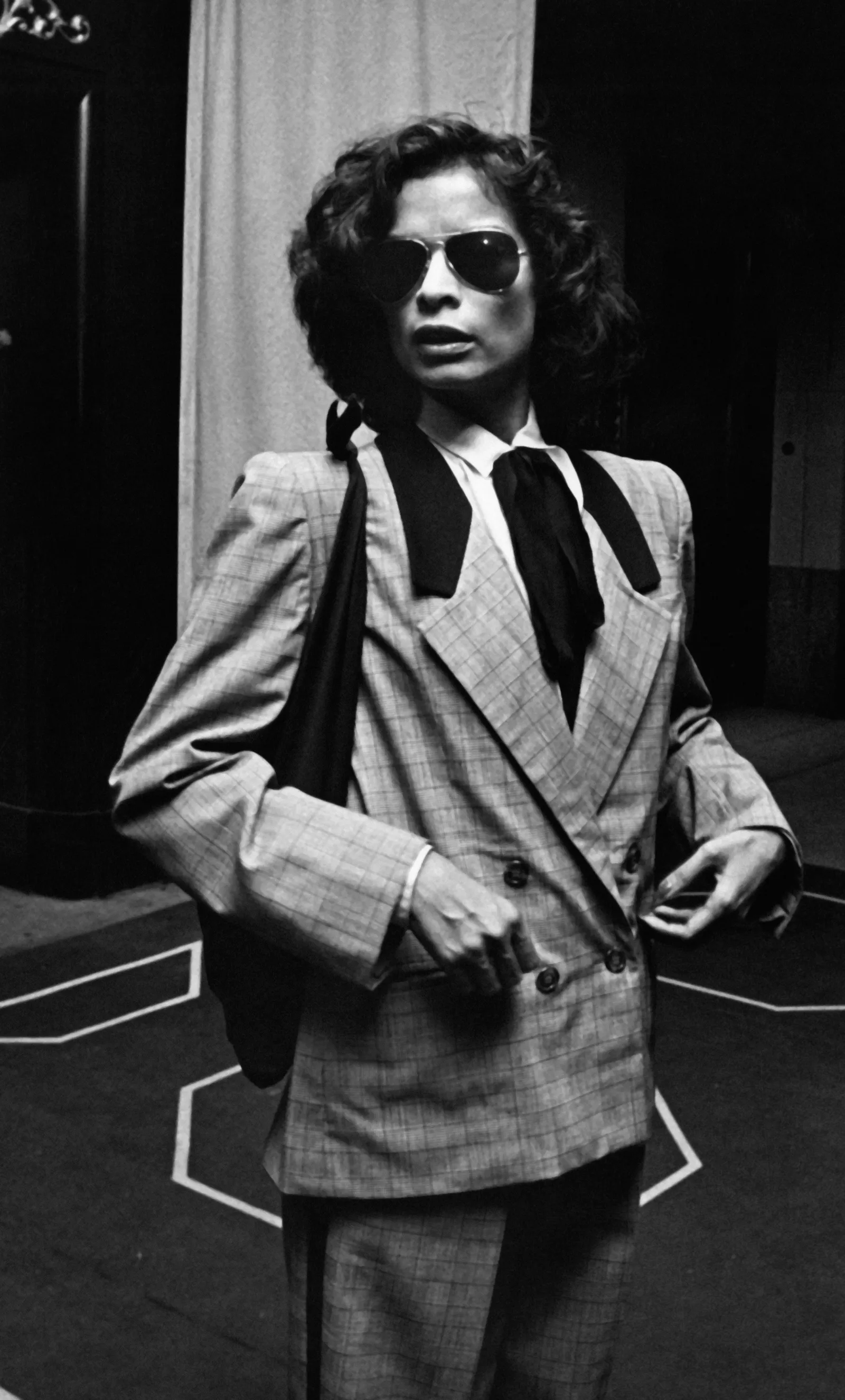 Bianca Jagger w eleganckim, dwurzędowym garniturze z ciemnymi okularami przeciwsłonecznymi.