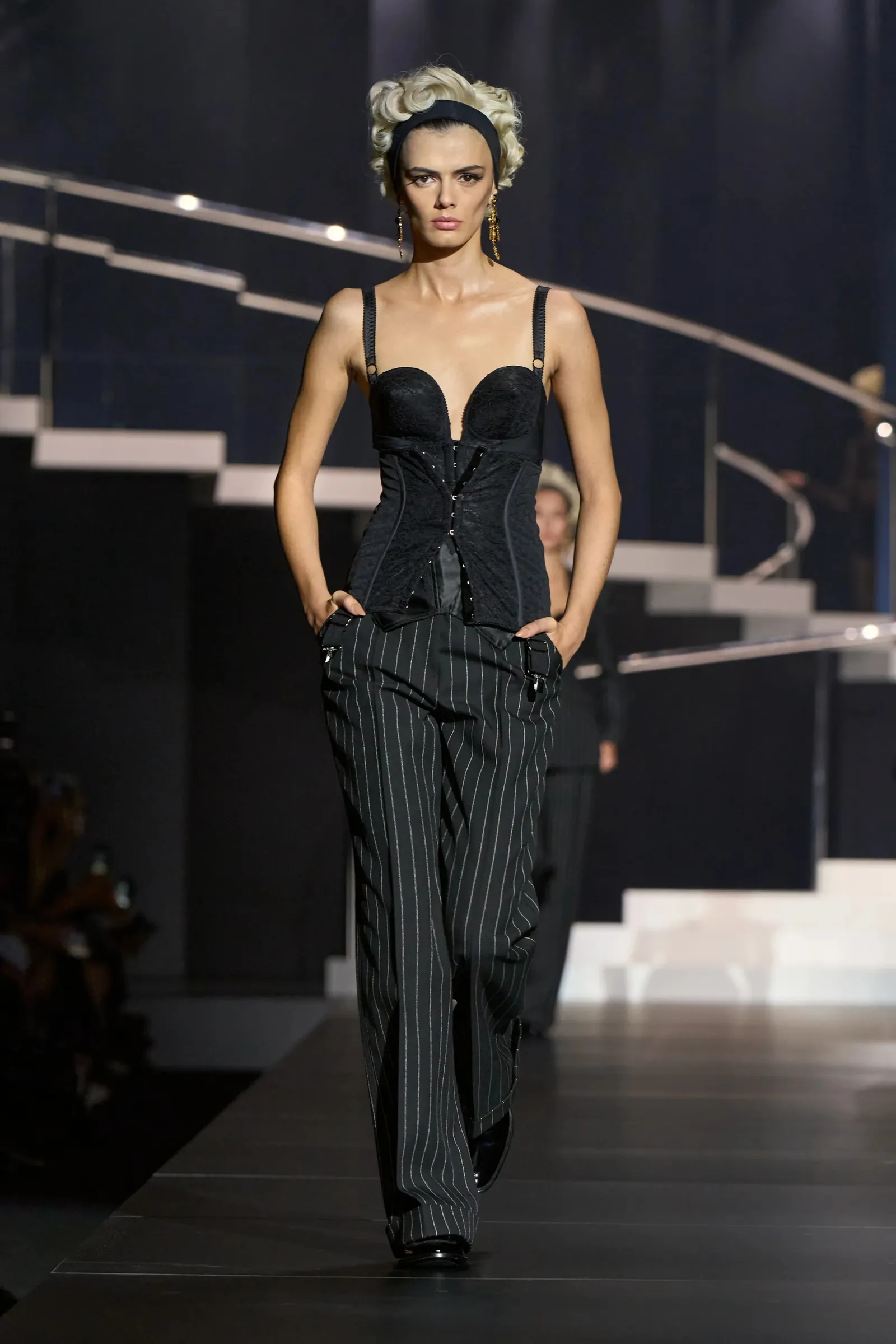 Modelka na wybiegu w eleganckim gorsecie oraz paskowanych spodniach, z charakterystycznym uczesaniem w stylu retro.