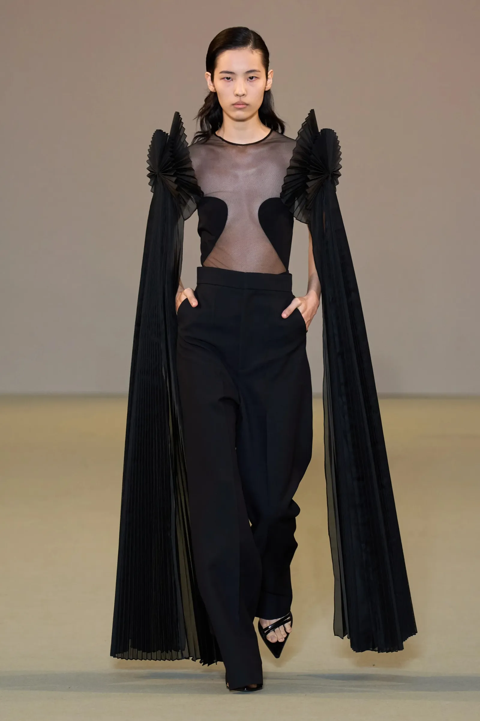 Modelka w eleganckiej czarnej kreacji z przezroczystą górą i długimi, plisowanymi rękawami, stoi na wybiegu.