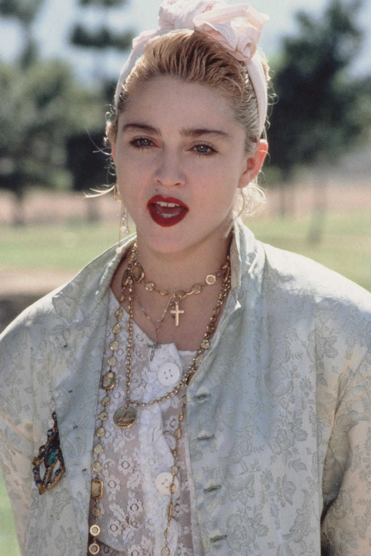 Madonna w stylizacji z jasną bluzką i długim naszyjnikiem, z różową opaską na głowie, na tle zieleni.