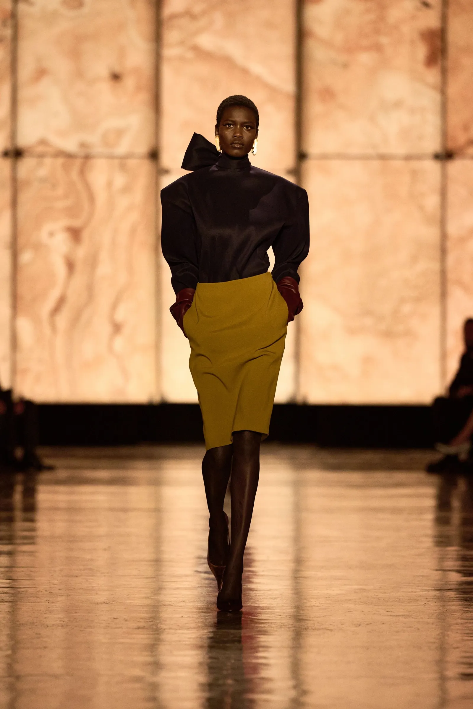 Modelka idzie po wybiegu w bluzce z kokardą YSL i zielonej spódnicy, prezentując nowoczesny styl mody.