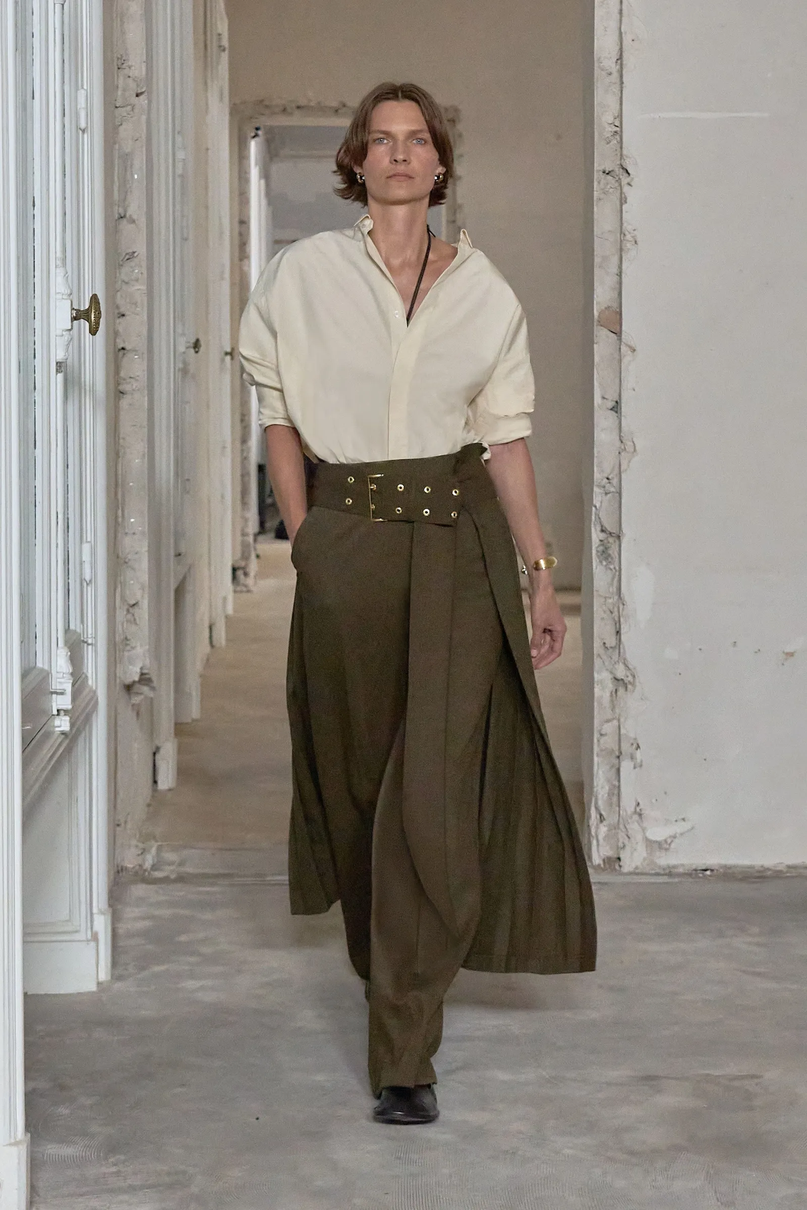 Model w brązowych spodniach i białej koszuli, idący przez przestronną, elegancką przestrzeń.