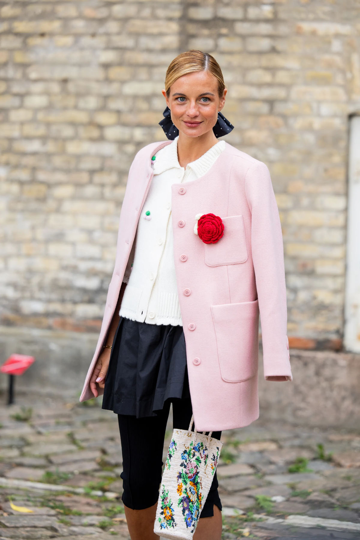 Kobieta w różowym płaszczu z broszką w kształcie czerwonej róży, w eleganckim stylu, z torebką haftowaną kwiatami.