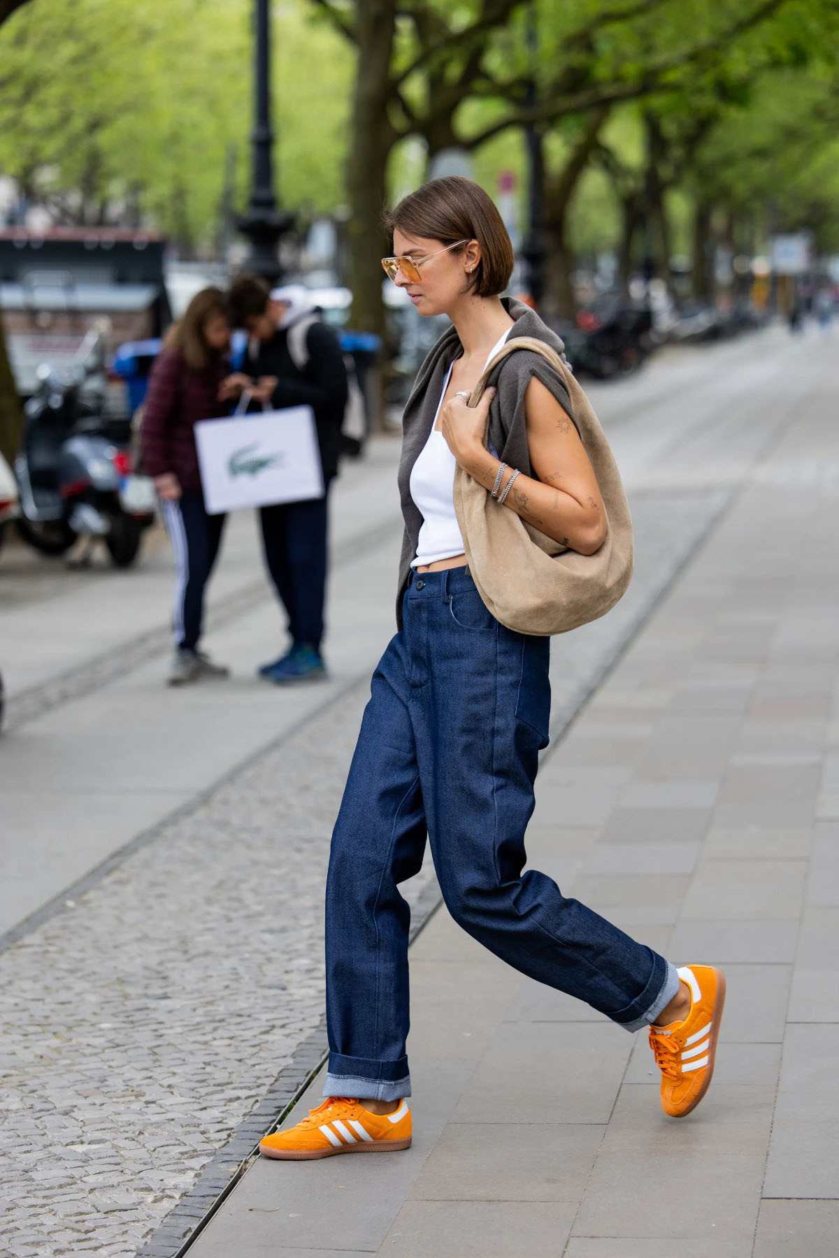 Kobieta w trampkach i cropped jeansach idzie ulicą, torba na ramieniu, otoczona zielenią drzew.