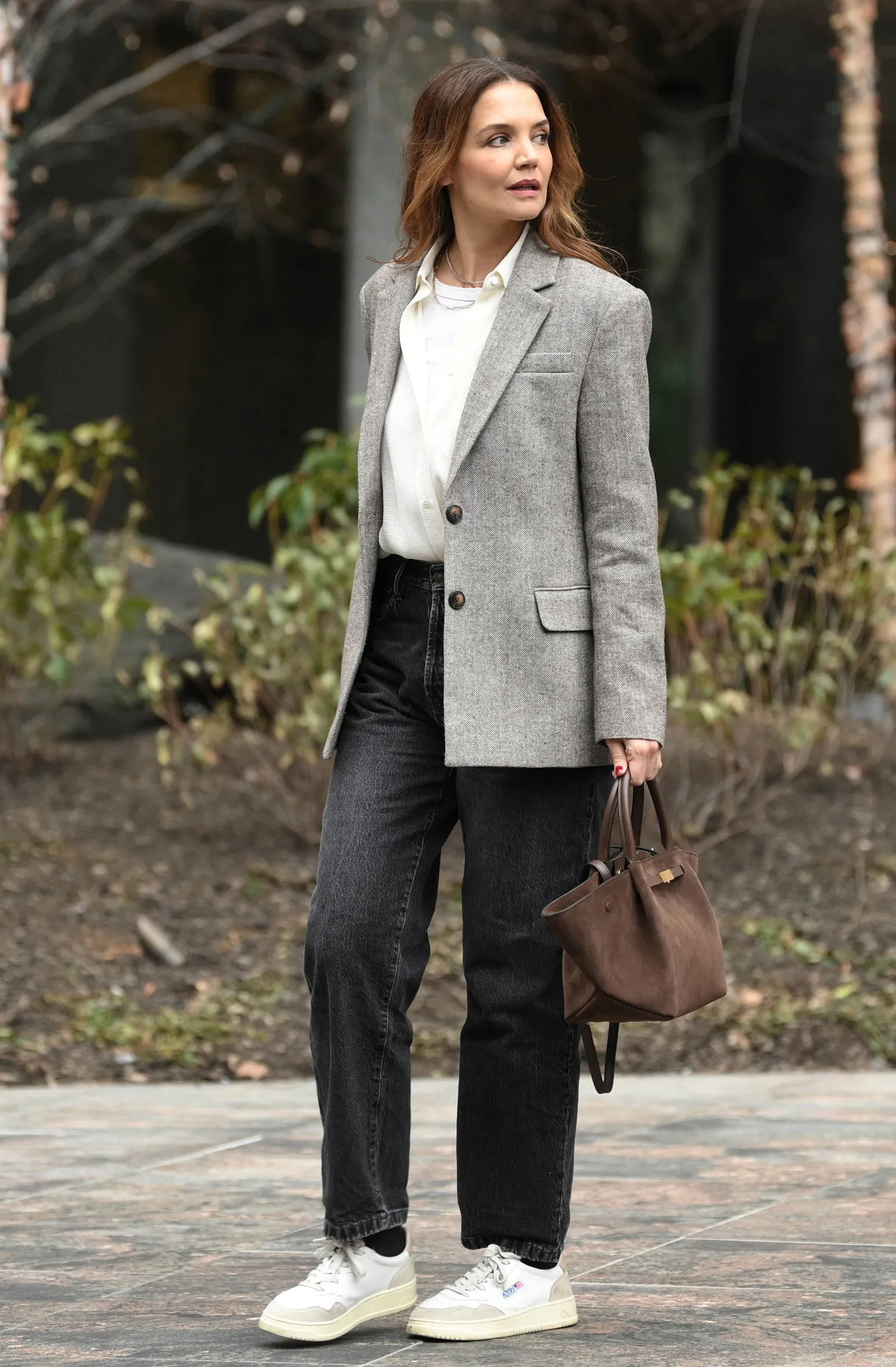 Katie Holmes w stylizacji z trampkami, cropped jeans i szarym żakietem, spaceruje w otoczeniu zieleni.