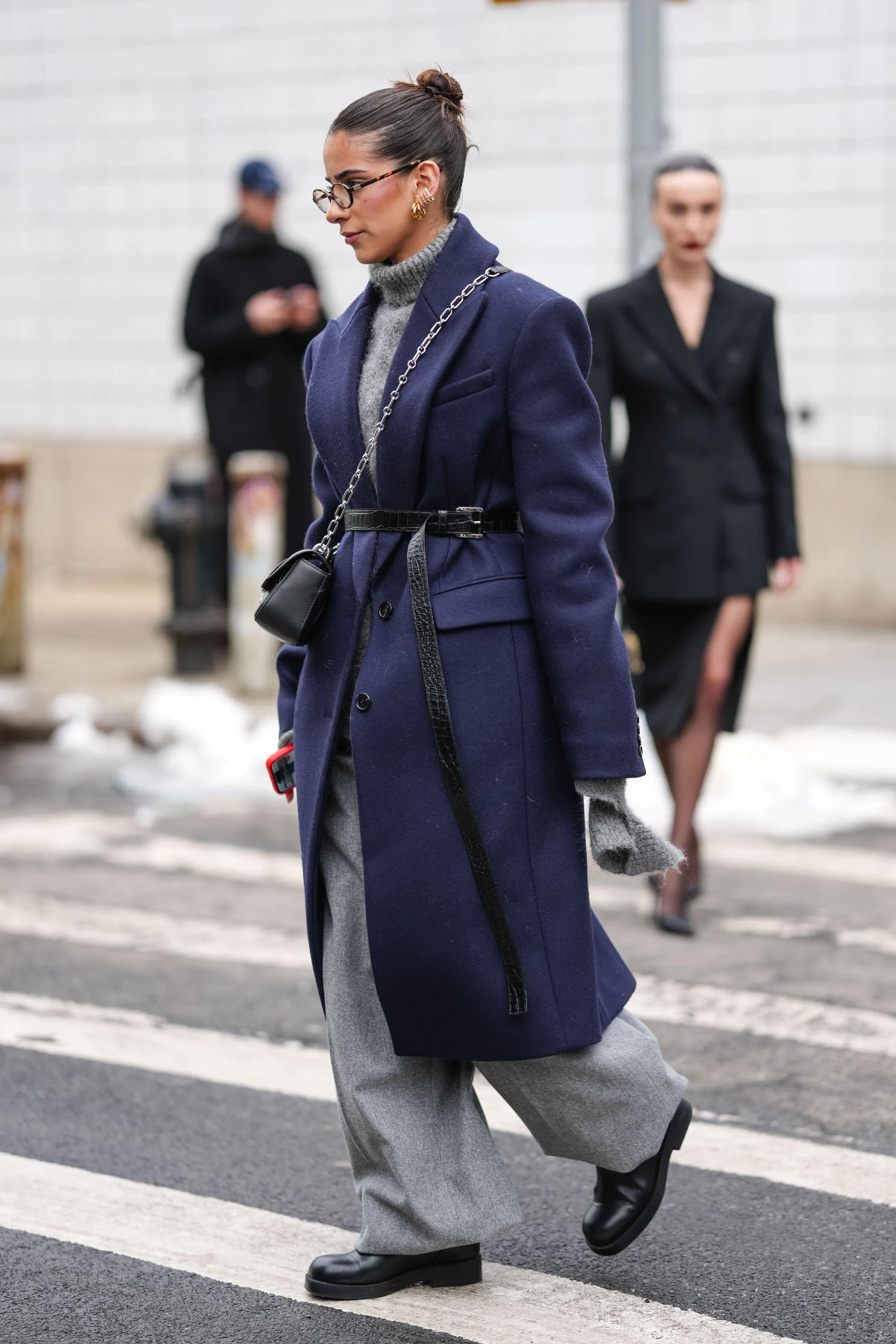 Stylowa osoba w granatowym płaszczu na zimę 2025 idzie przez jezdnię, nosząc szare spodnie i torebkę.