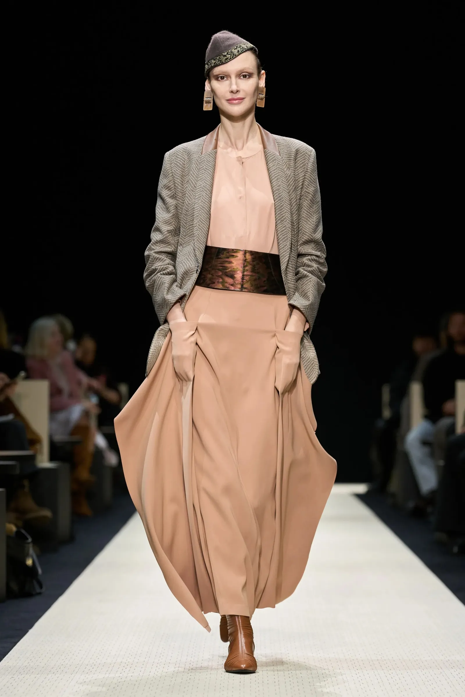 Modelka w beżowej sukience i marynarce prezentuje elegancki outfit na wybiegu.
