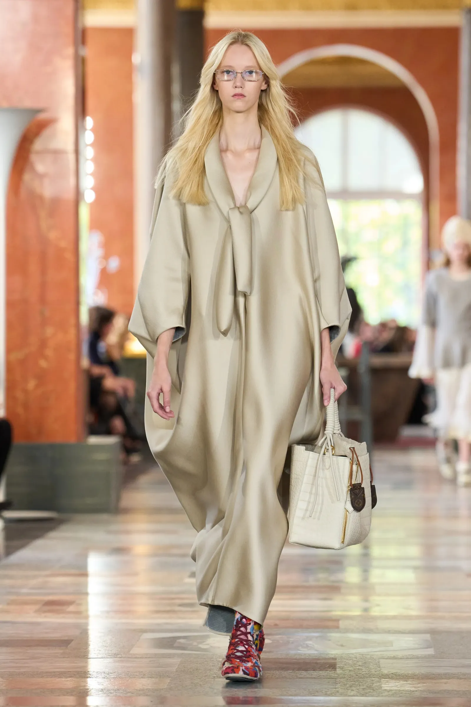 Modelka w długim, beżowym płaszczu, kolorowe botki Louis Vuitton Jesień-Zima 2025, z torebką w dłoni.