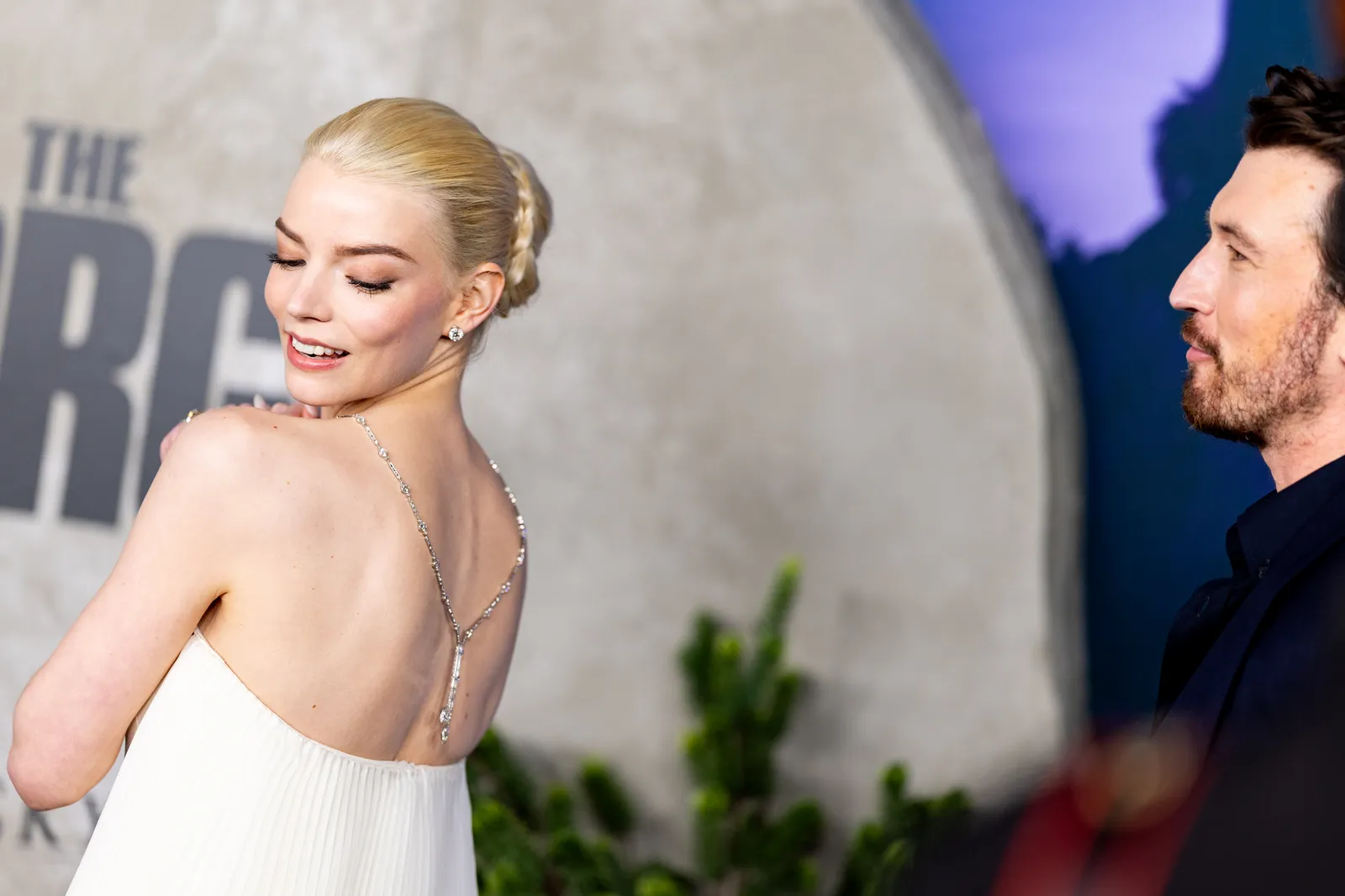 Anya Taylor-Joy w białej sukience z naszyjnikiem na plecach uśmiecha się, a mężczyzna z zainteresowaniem ją obserwuje.