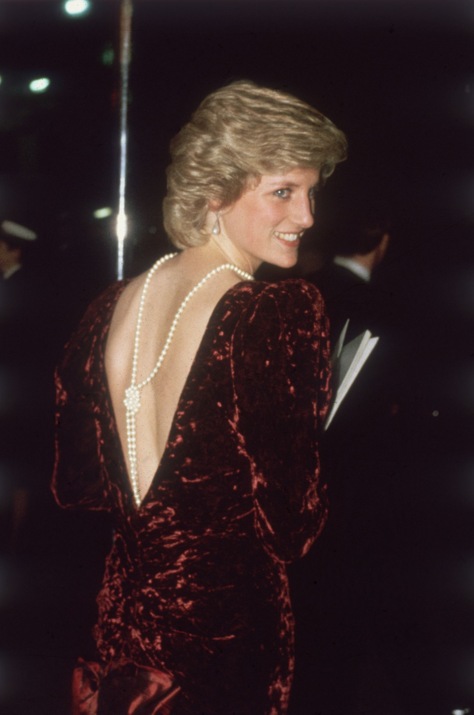 Lady Di w burgundowej sukni z głębokim dekoltem i pięknym naszyjnikiem na plecach, uśmiecha się do fotografii.