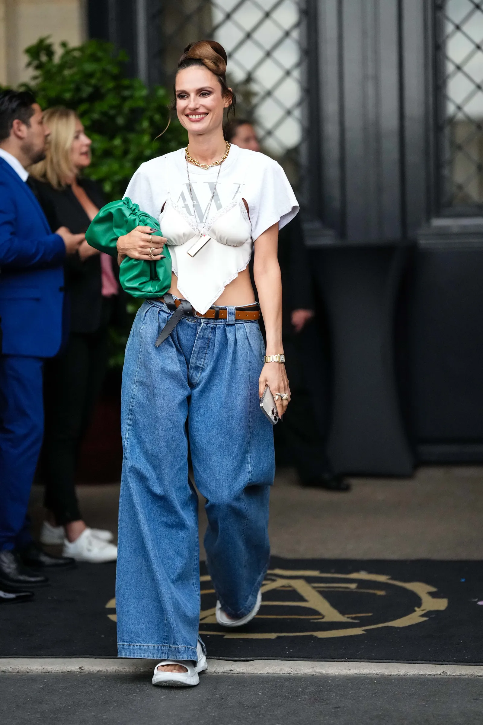 Kobieta w baggy jeans i biała bluzka, uśmiechnięta, przechodzi obok grupy ludzi.