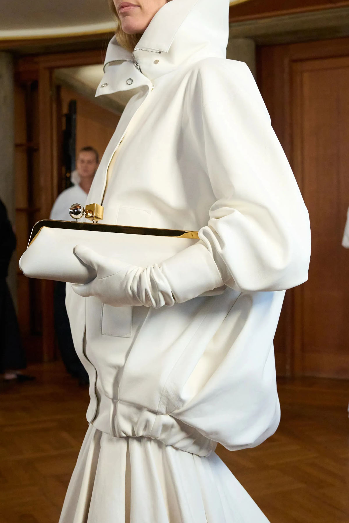 Modelka w białej, eleganckiej odzieży trzyma torebkę na zatrzask marki Jacquemus.