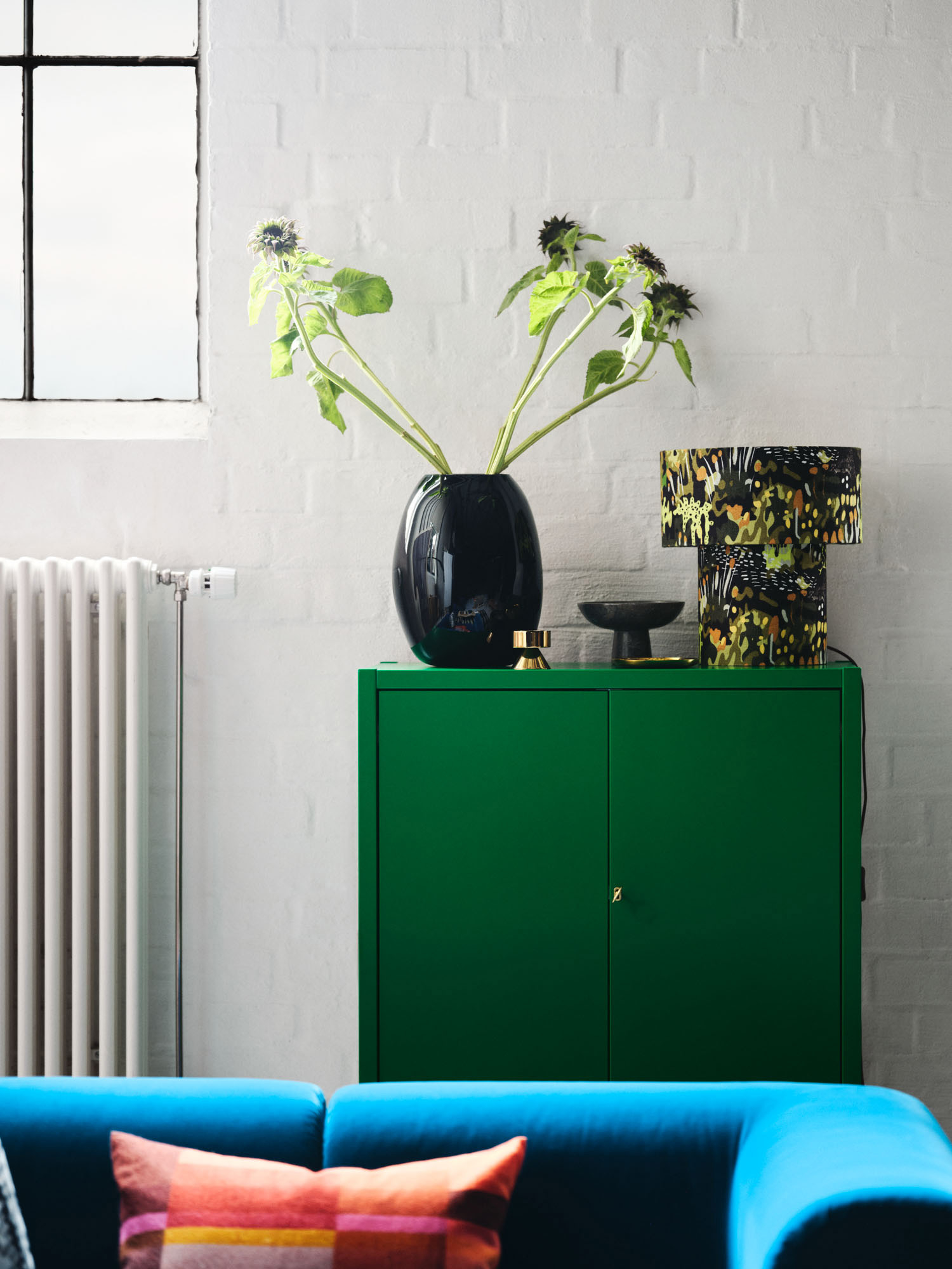 Wnętrze z zieloną szafką Ikea, czarnym wazonem i kolorową lampą, oraz niebieską kanapą z poduszkami.
