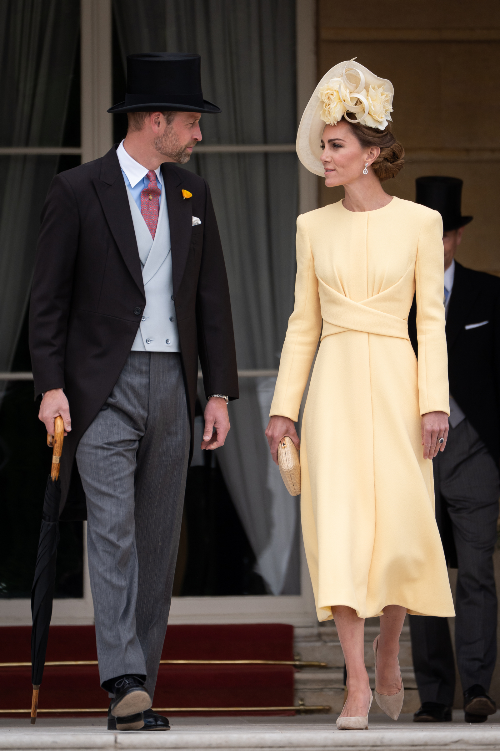 Księżna Kate w maślanym żółtym stroju, spaceruje z mężczyzną w eleganckim garniturze i cylinderze.
