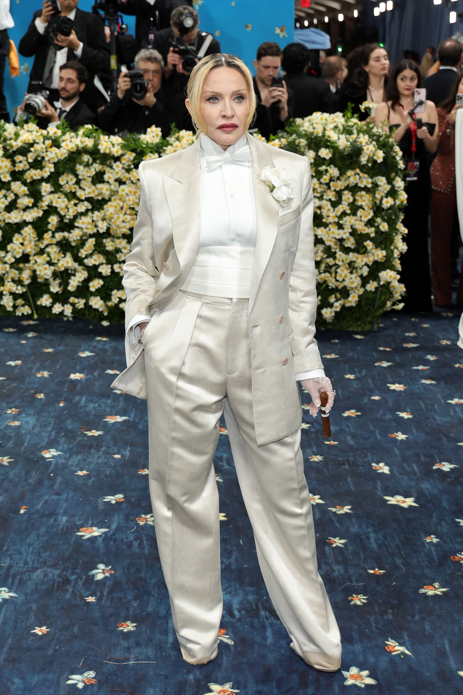 Madonna w białym smokingu, prezentująca elegancki styl na tle kwiatowej dekoracji.