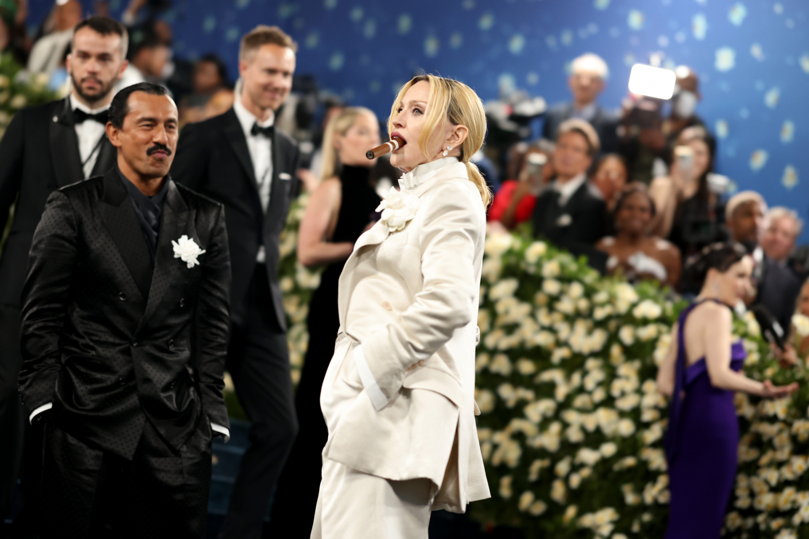Madonna w białym smokingu na scenie w towarzystwie innych elegancko ubranych osób, otoczona kwiatami.