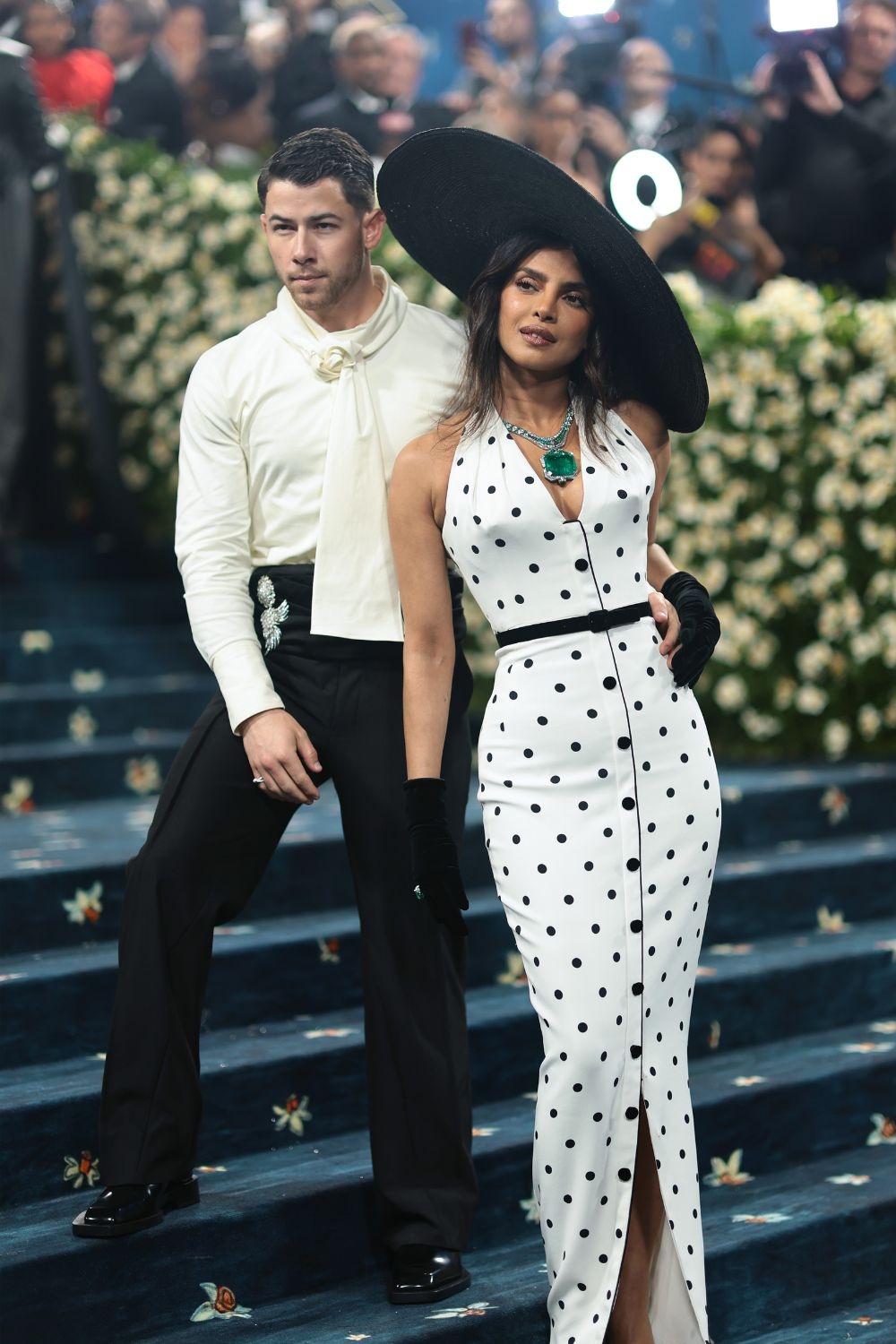Nick Jonas i Priyanka Chopra na met gala 2025, elegancko ubrani, wśród floralnych dekoracji, pozują na schodach.