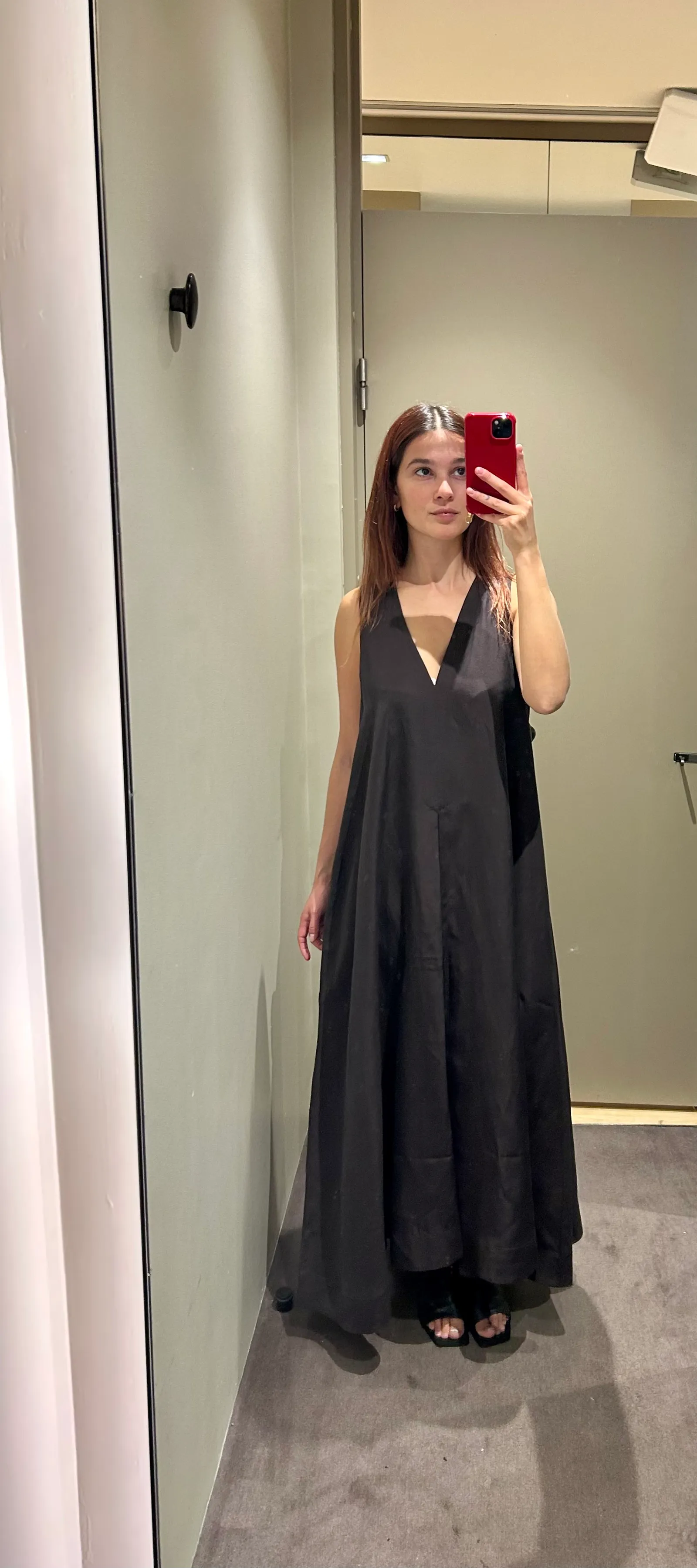 Kobieta w czarnej sukience na wesele stoi w przymierzalni, trzymając telefon. Odbicie w lustrze widoczne z boku.