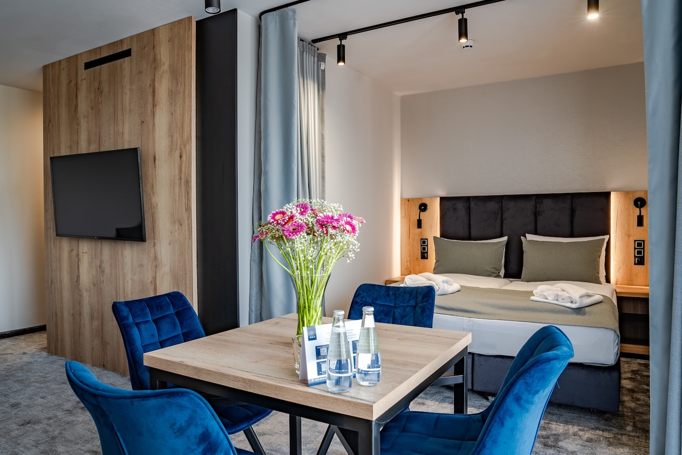 Nowoczesny pokój hotelowy Aqua by Grano w Szklarskiej Porębie z podwójnym łóżkiem, telewizorem oraz stołem z niebieskimi krzesłami i bukietem kwiatów.