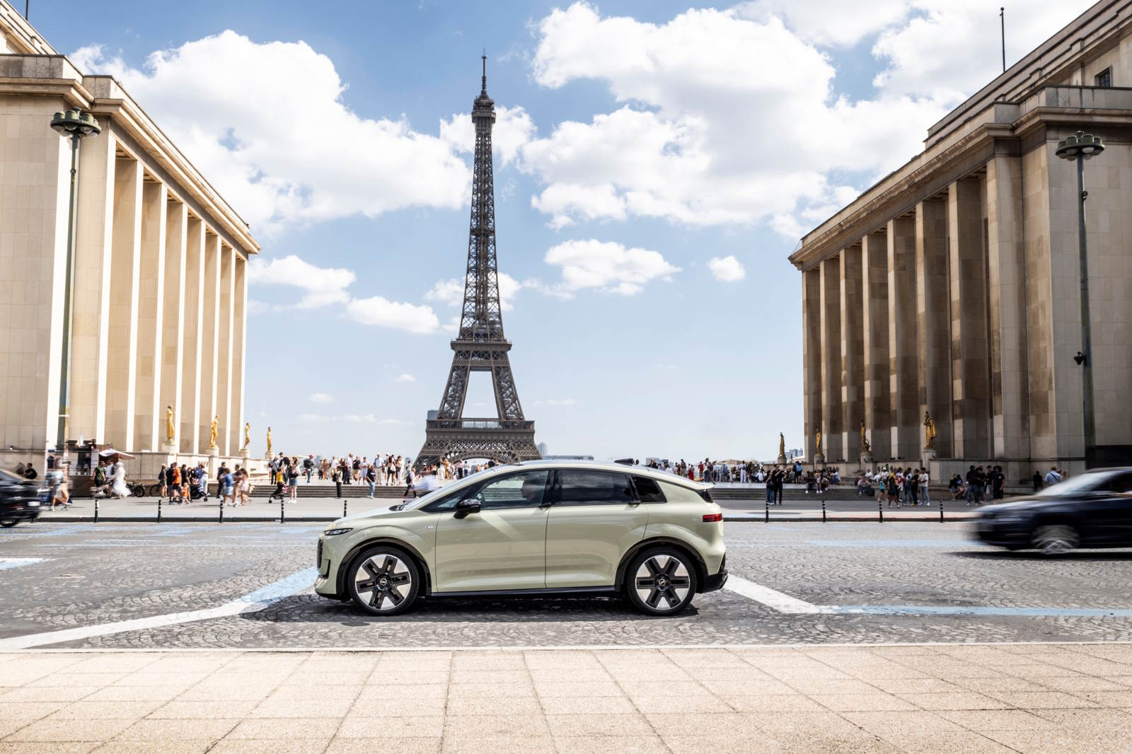 Nowoczesny pojazd osobowy Audi A2 e-tron w jasnym kolorze, stojący na tle Wieży Eiffla i tłumu turyst&oacute;w.