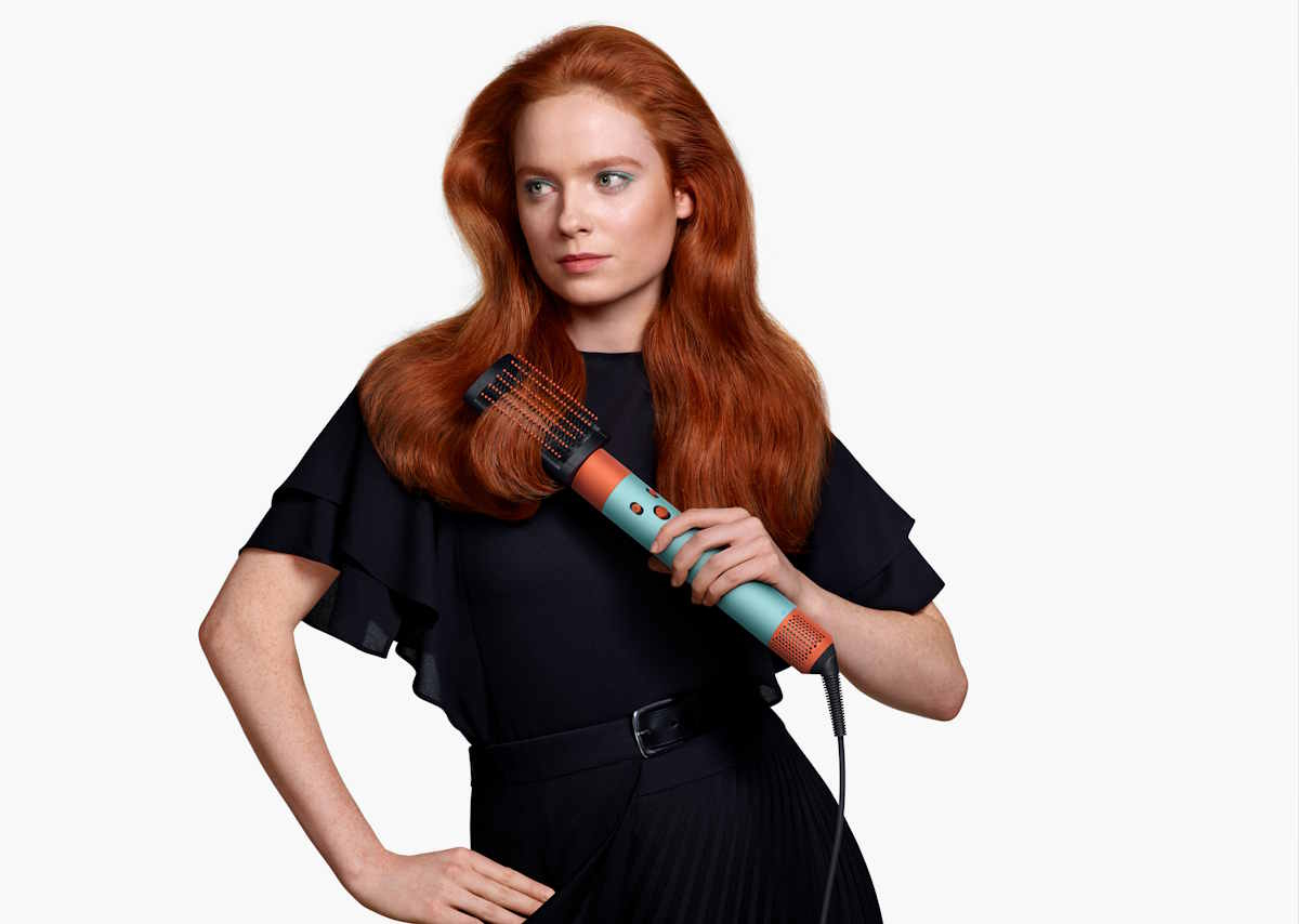 Kobieta z długimi rudymi włosami trzyma w ręku urządzenie do stylizacji włosów, nowy multistyler Dyson Airwrap i.d., ubrana w czarną bluzkę i spódnicę.