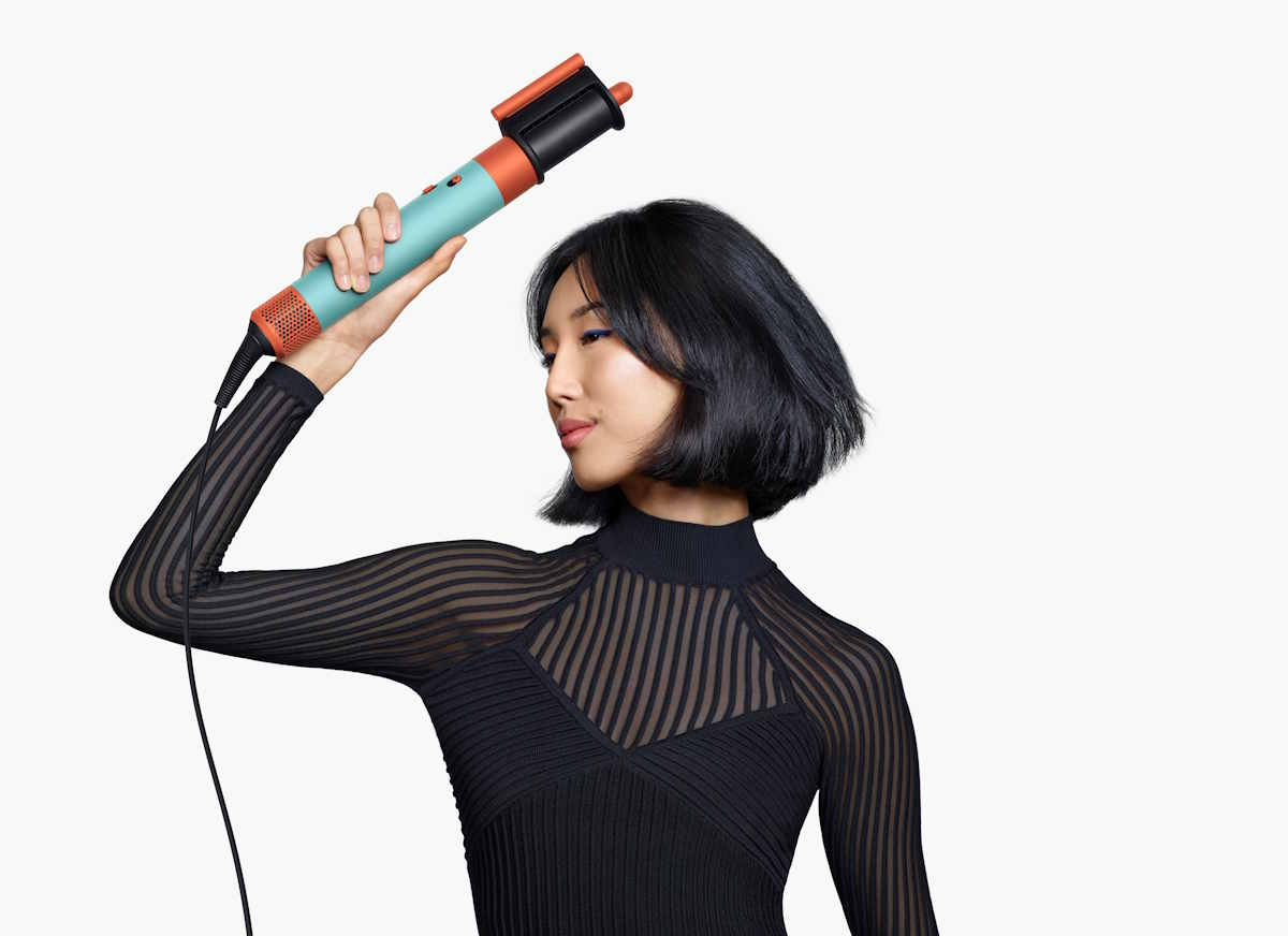 Kobieta w czarnym swetrze z prześwitującymi detalami trzyma nowy multistyler Dyson Airwrap i.d. Ma krótkie, ciemne włosy i uśmiecha się.