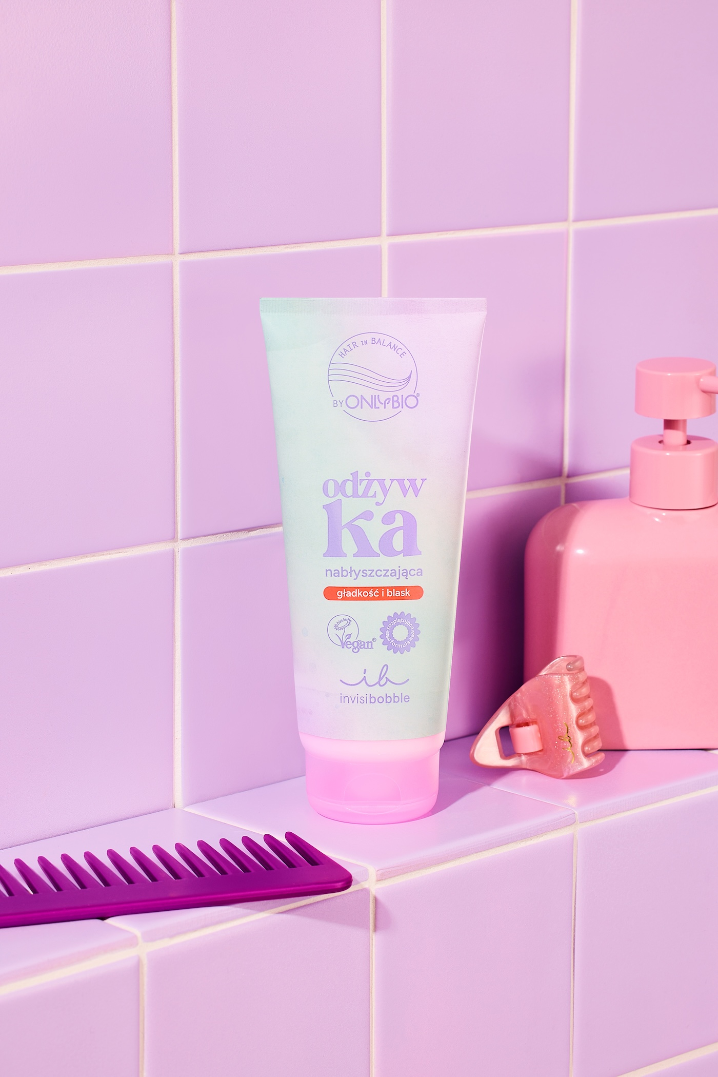 Odżywka nabłyszczająca OnlyBio x invisibobble, w tle fioletowe płytki łazienkowe, różowa spinka do włosów, różowe opakowanie z dozownikiem i purpurowy grzebień.